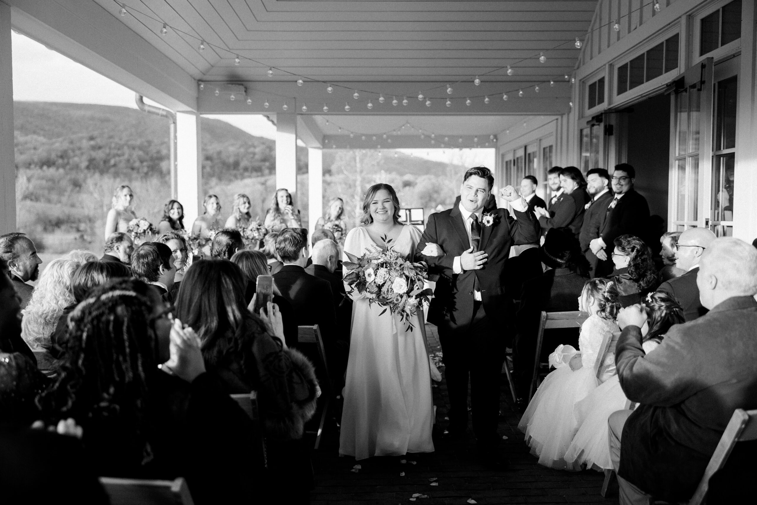 Black and white image of bride and groom happily walking back up the aisle, groom's fist in the air as they celebrate their king family vinyeards fall wedding