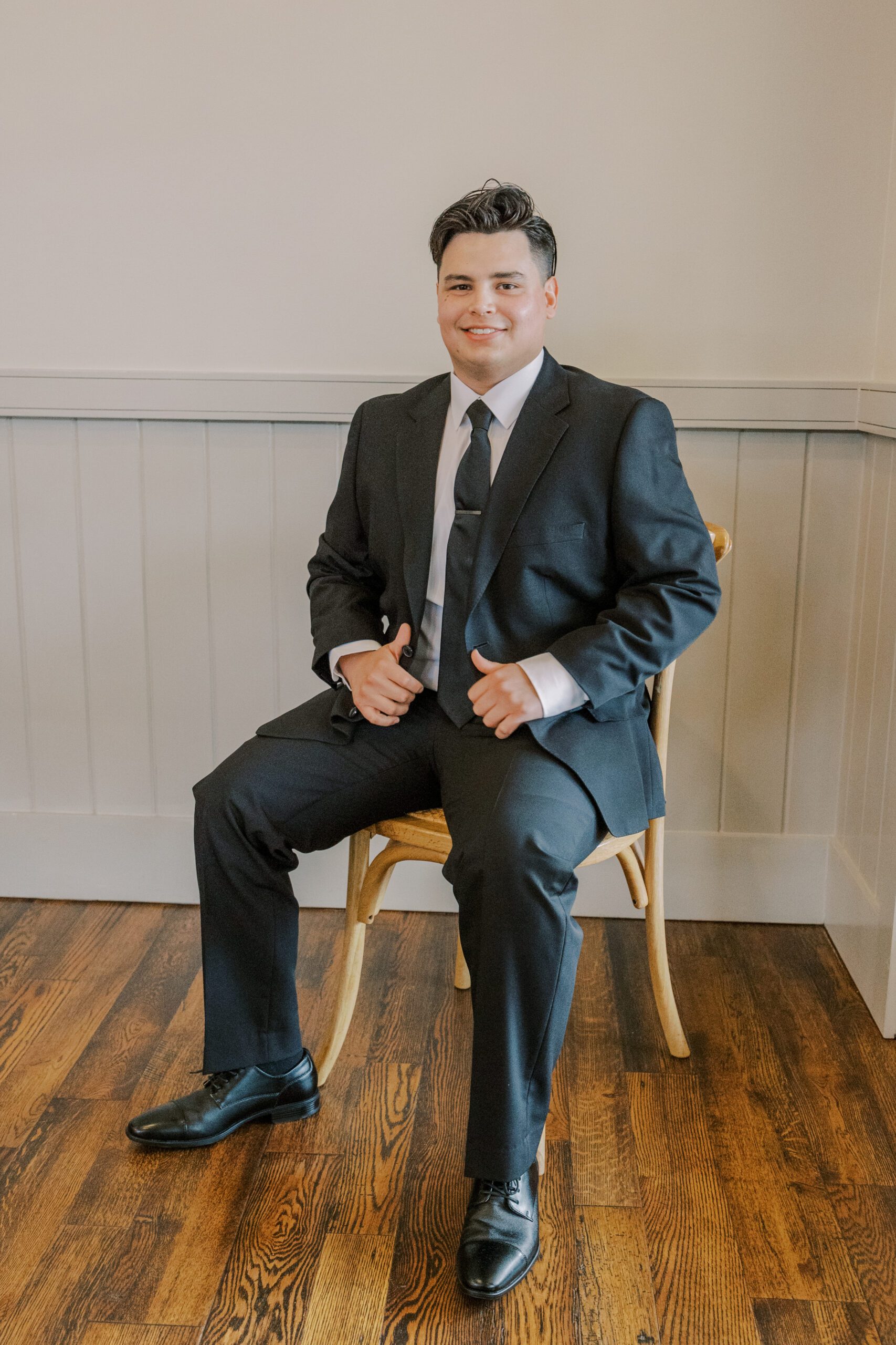 Groom sitting on chair sitting while wearing his suit smiling at camera