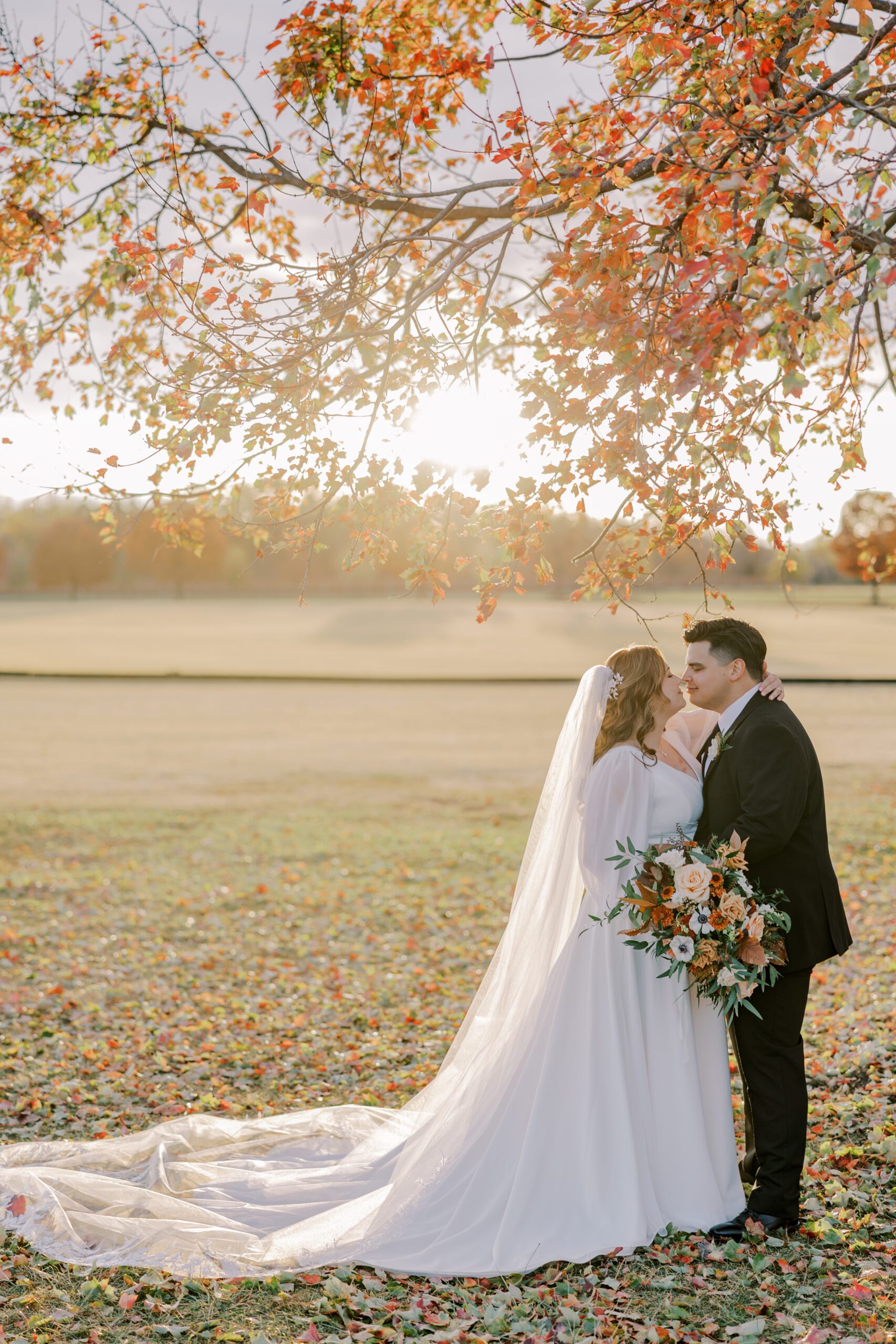 Beautiful full length sunset image of bride and groom touching noses while standing under a tree with orange leaves on it at king family vineyards