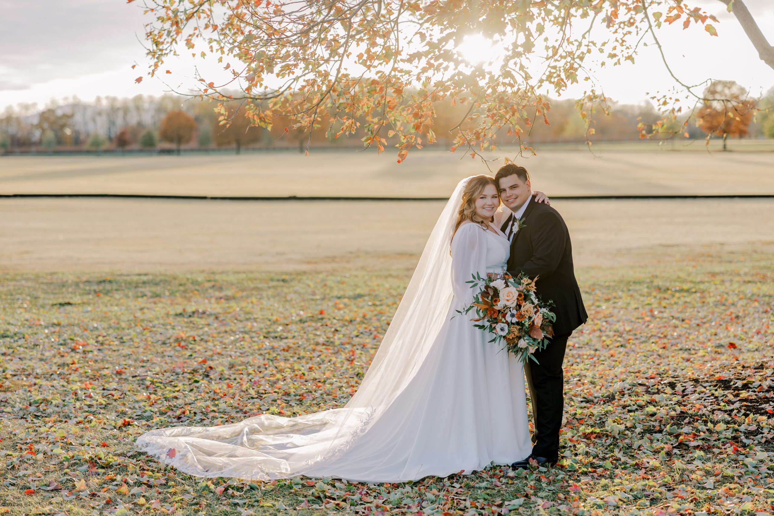 Bride and groom both smiling at camera standing under a tree with orange leaves as the sun begins to set