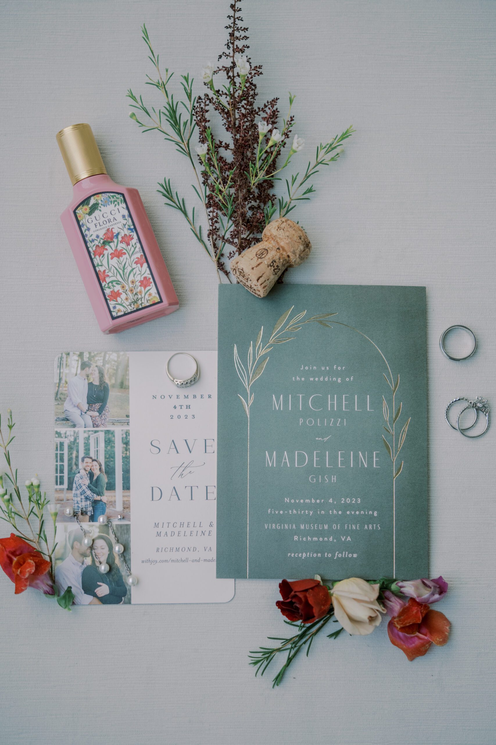 Detail photo of the couple's save the date card and wedding invitation, also pictured are the couple's rings, a perfume, cork, and some flowers