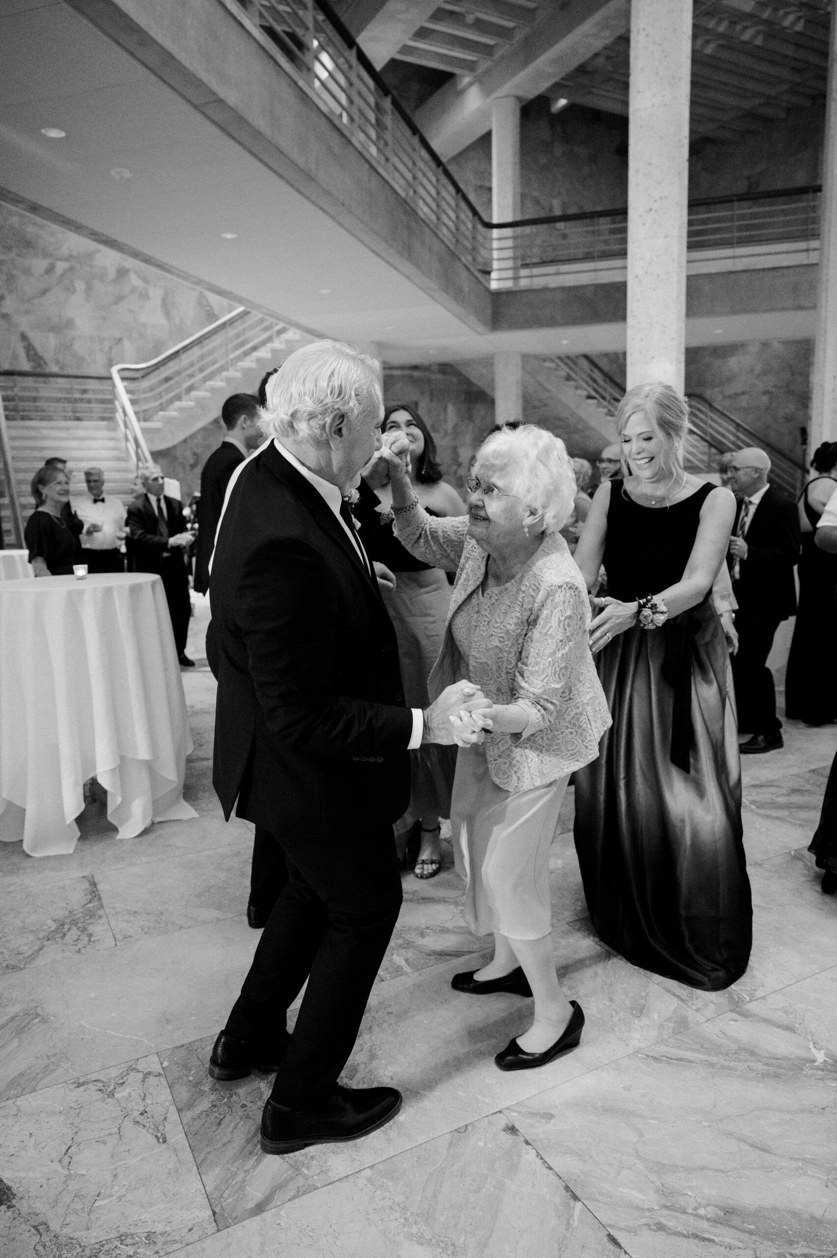 Black and white photo of older couple dancing, mother of bride stands behind the woman to gently support her while she dances
