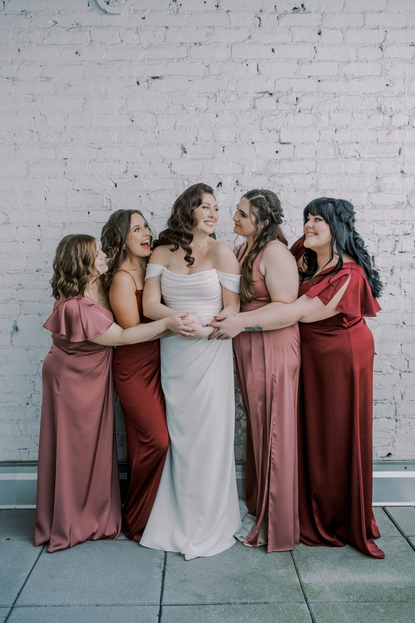 Full length photo of bride in her white wedding dress with 2 bridesmaids on each side of her alternating red and rose colored dresses, all smiling at one another