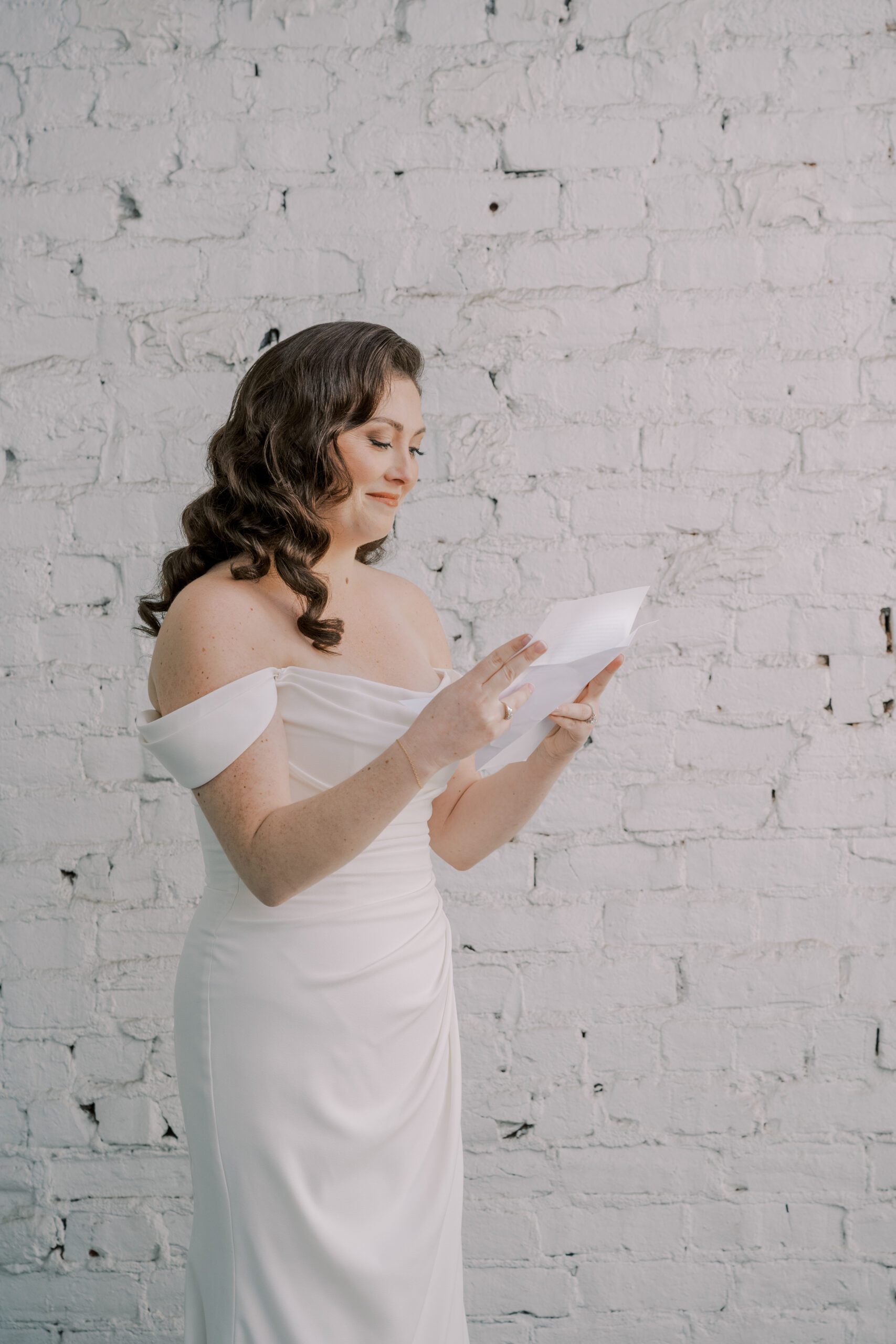Bride in her dress smiling at a letter she is reading in front of a white brick wall at vmfa