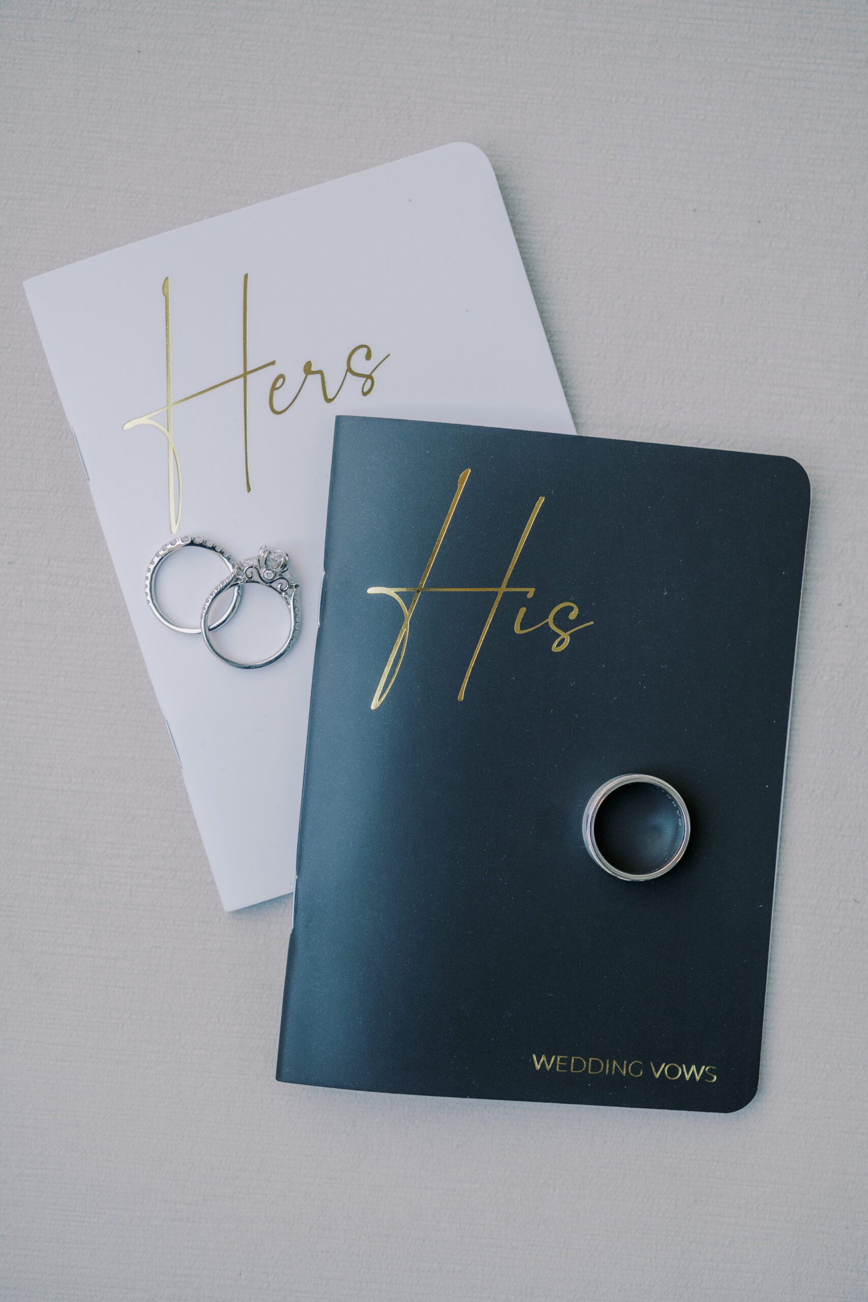 Image of wedding vow notebooks, a black one that says his in gold and a white one that says hers in gold, also pictured are the couple's rings
