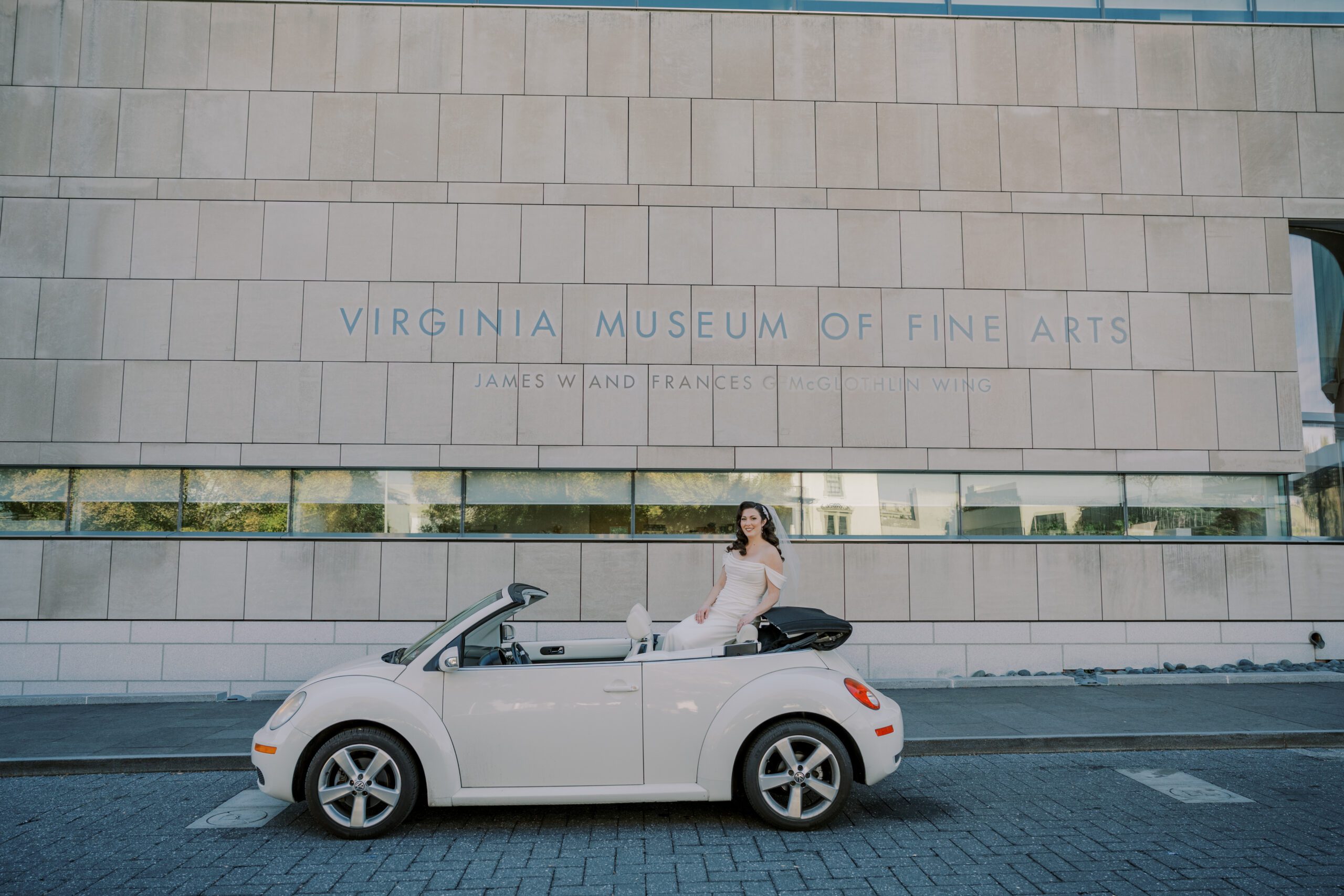 Bride sitting up on the back of a convertible white volkswagen beetle car in front of the virginia museum of fine arts