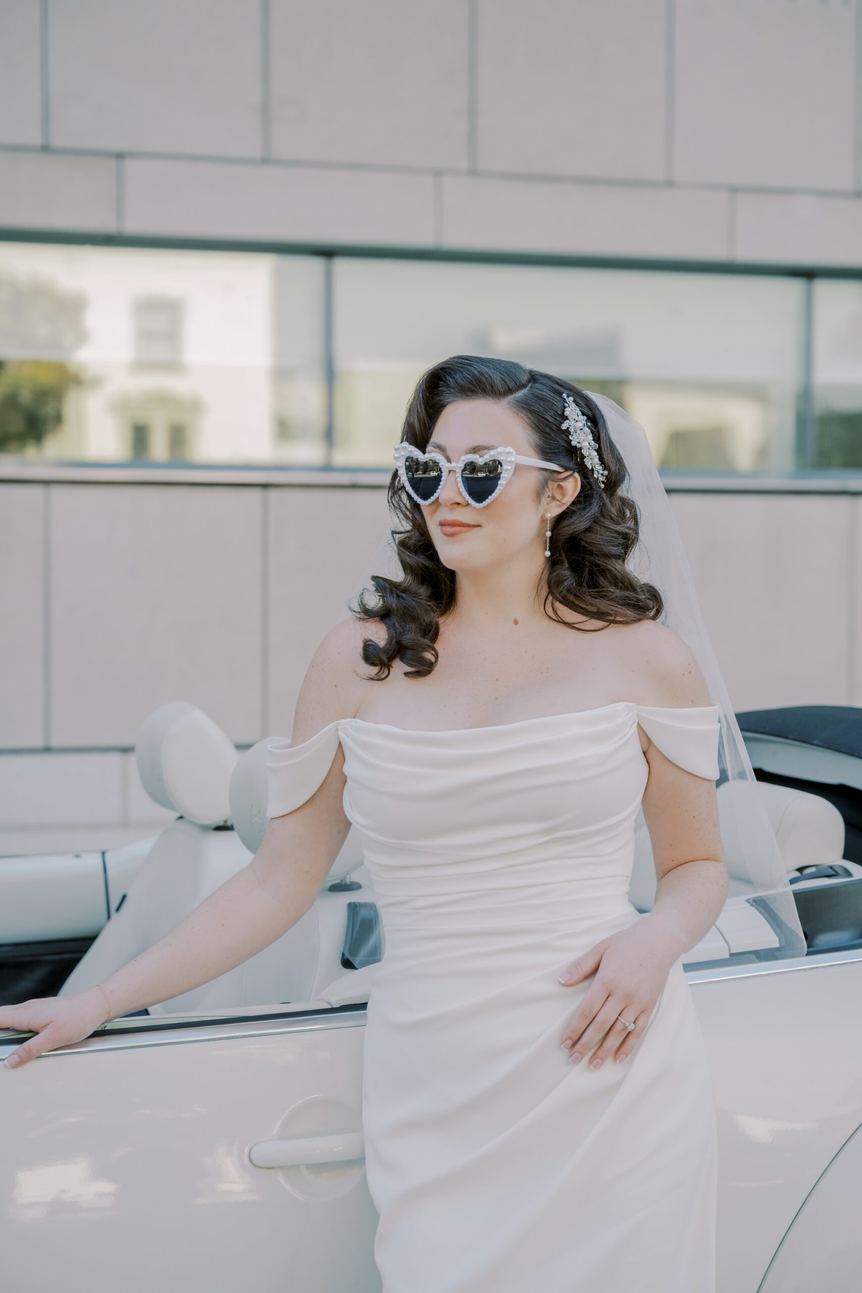 Bride standing in front of white convertible car looking off into the distance wearing white heart shaped sunglasses, her wedding dress and veil