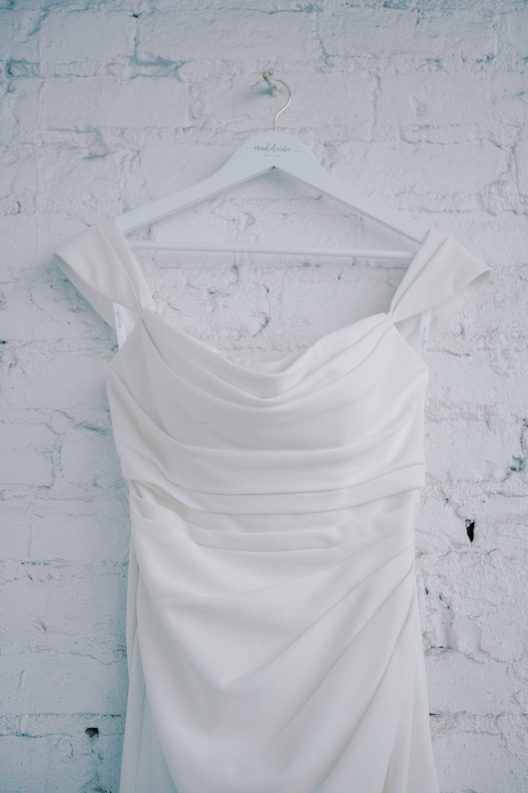 Waist up photo of a white wedding dress hanging from a white hanger against a white brick wall