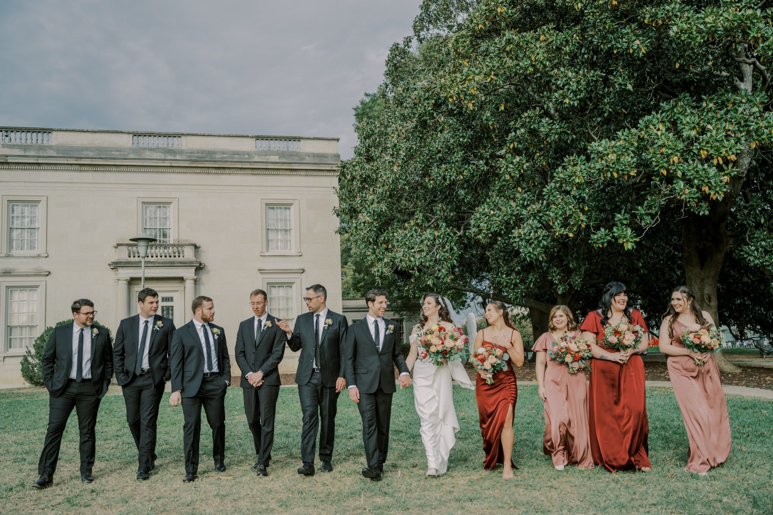 Bride and groom walking in grass with their groomsmen and bridesmaids on either sides of them, all looking at one another smiling and chatting