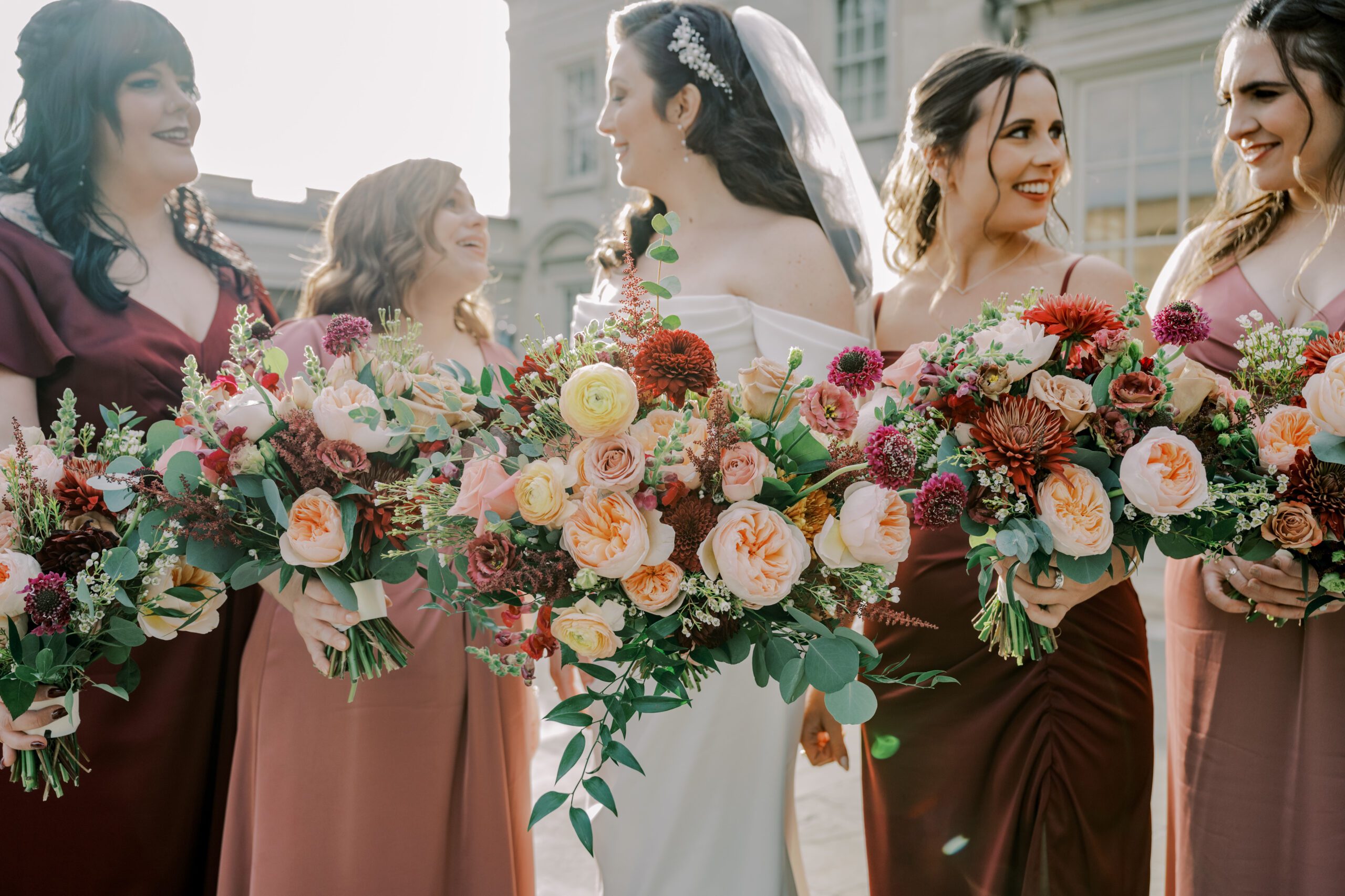 Close up image of floral bouquets with red, pink, burgundy, and peach colored flowers, bride and bridesmaids slightly out of focus in background looking at one another
