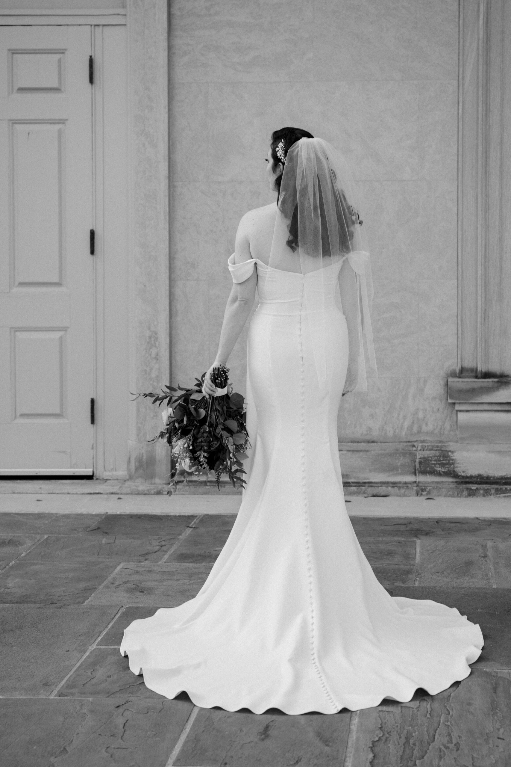 Black and white image of bride facing away from camera, buttons run down the back of her dress, and a veil hanging from her head, she has her arms down and in one hand has her bouquet facing down.