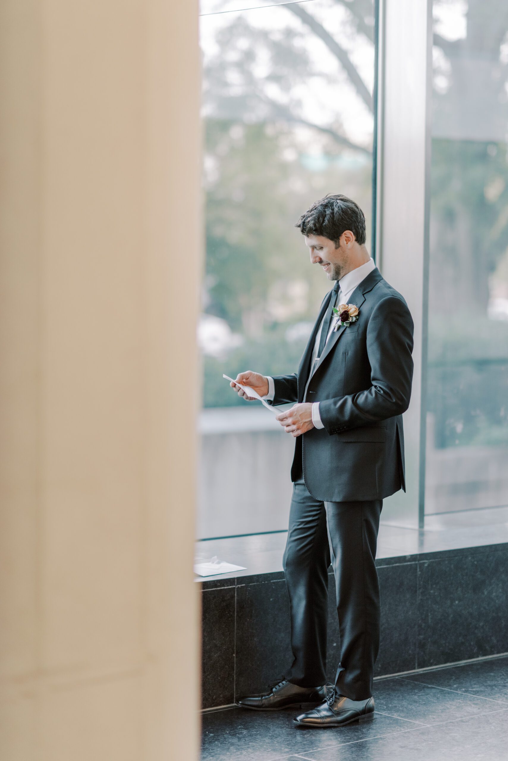 Photo of groom standing, taken from afar as he smiles down at a letter he is reading