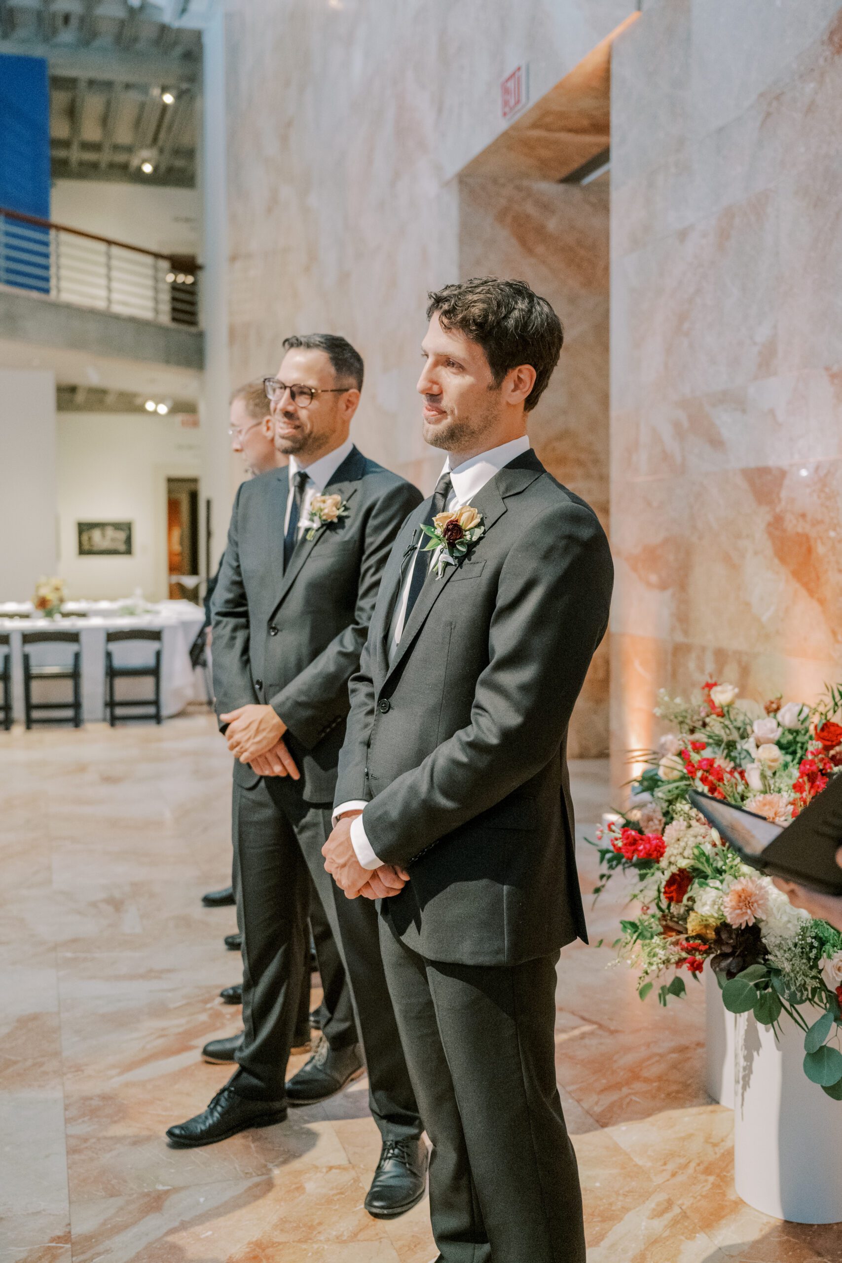 Groom standing at alter looking towards where bride would be, his groomsmen pictured in background, pink toned marble walls and floors inside vmfa