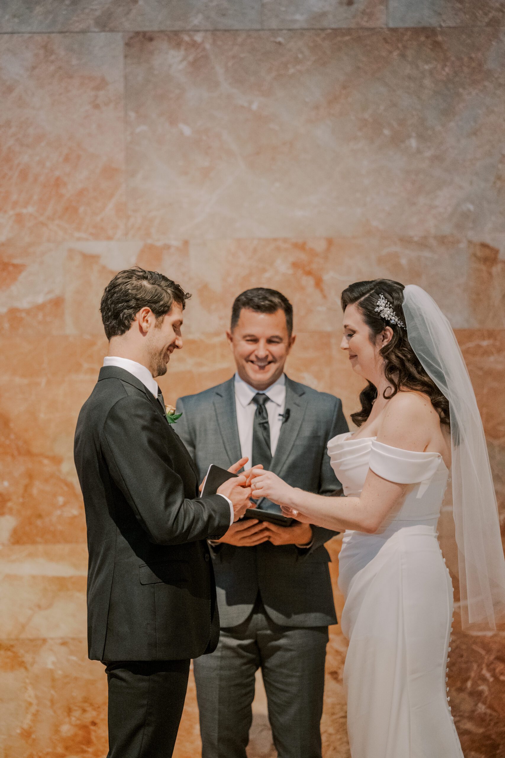 Bride and groom exchanging rings during their ceremony, smiling, with their officiant behind them and a pink marble wall in backgound