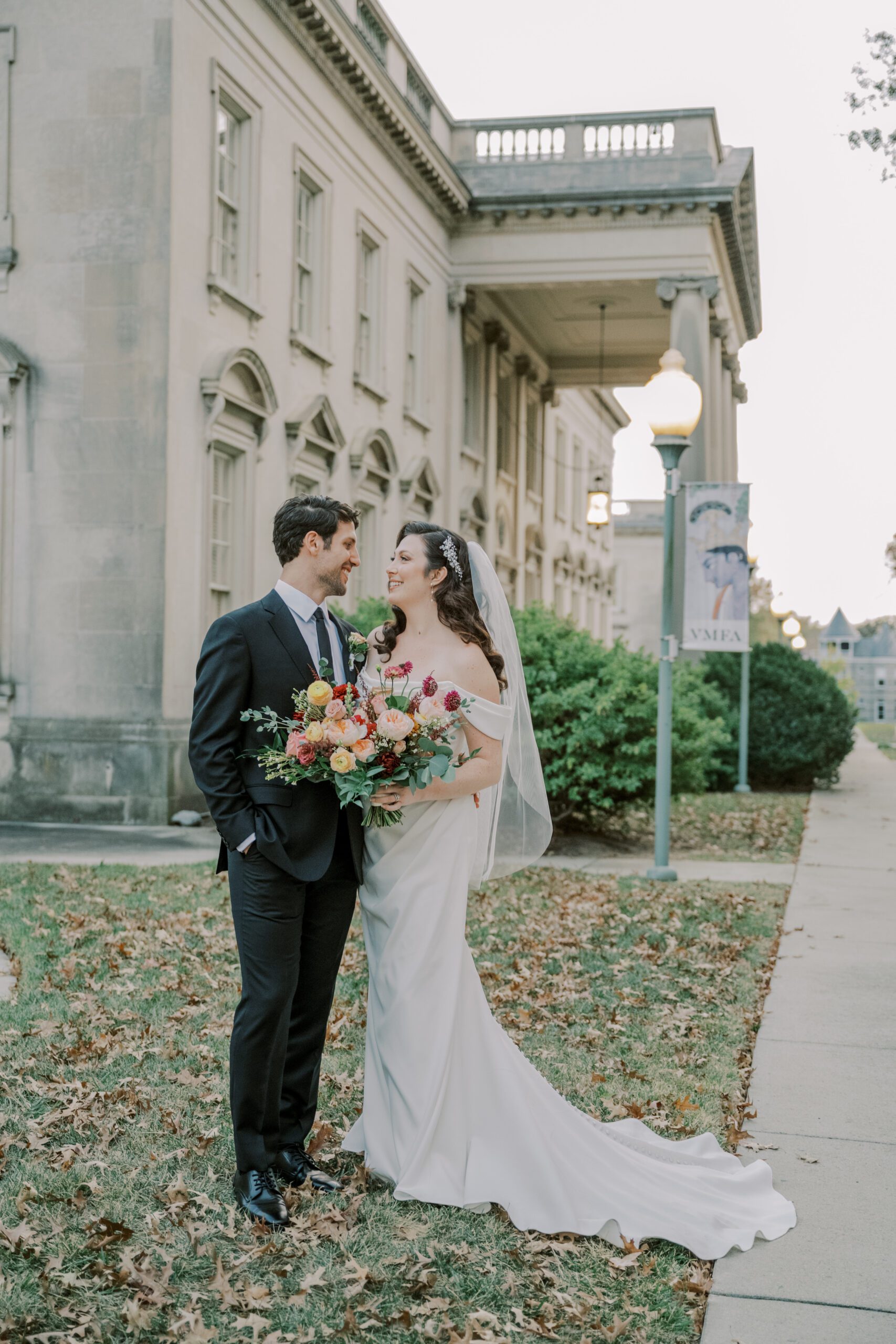 Outdoor full length photo of bride and groom smiling at one another while bride holds her bouquet in front of vmfa