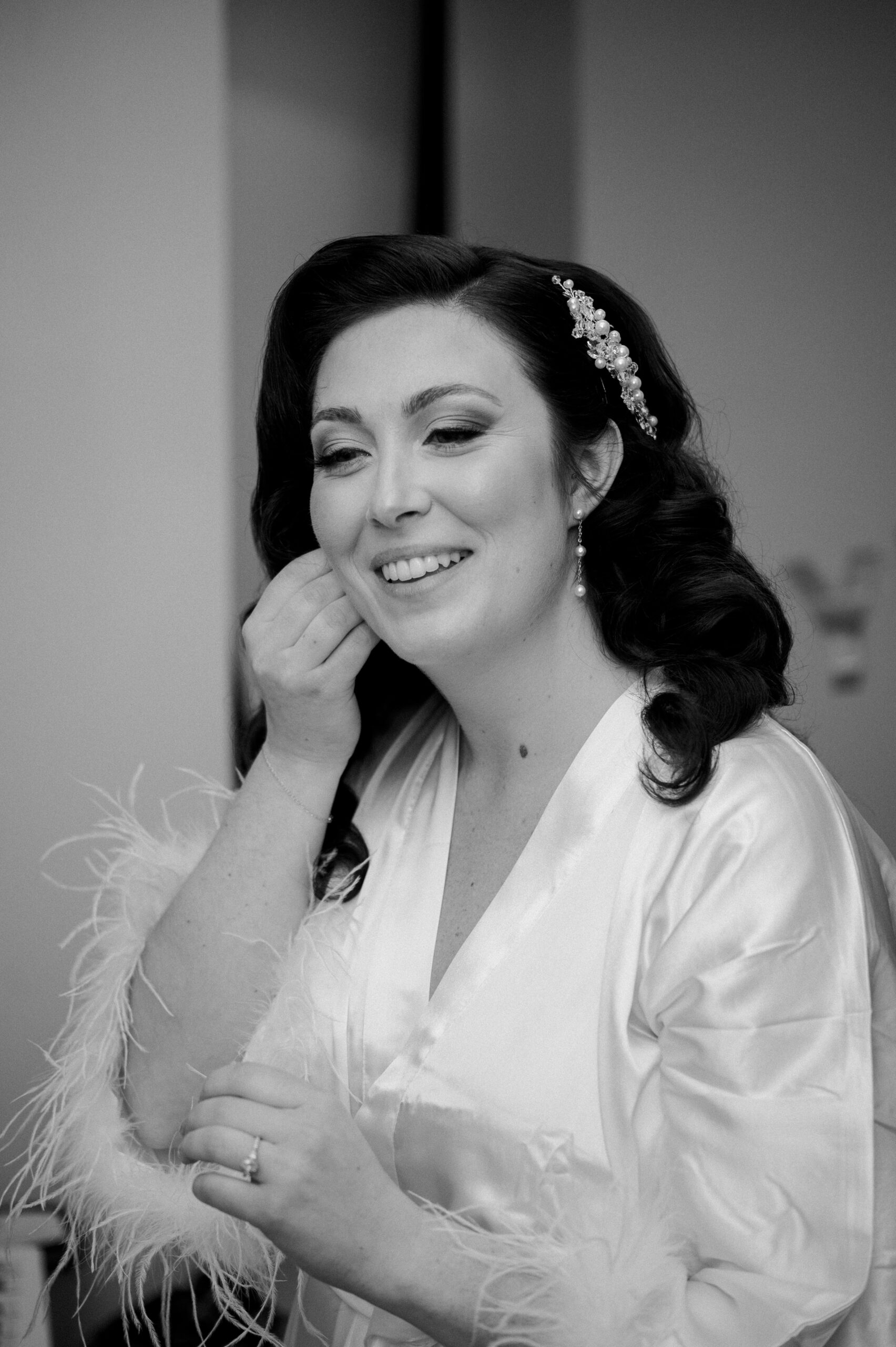 black and white photo of bride with her hair and makeup done, putting in one of her earrings while wearing a white robe with feathered details at end of sleeves