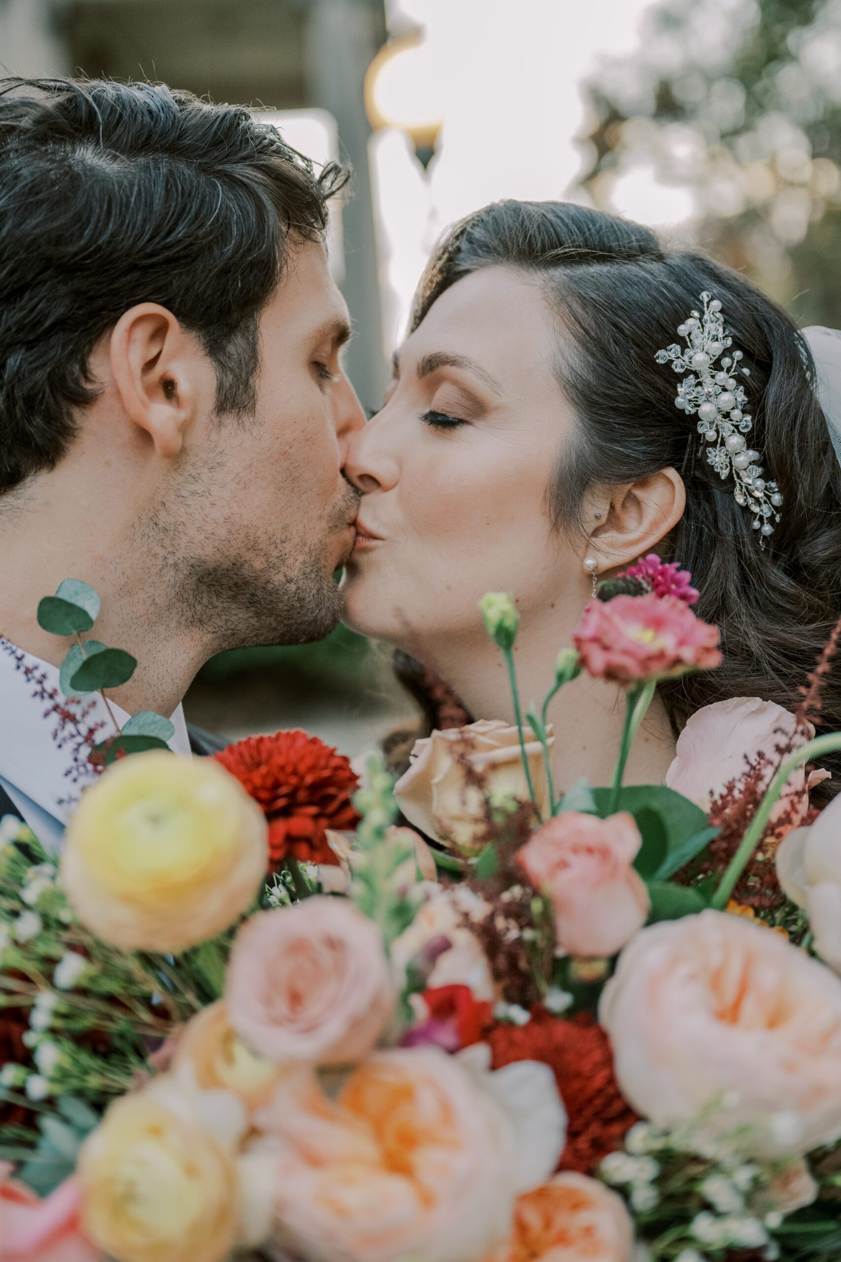 Close up image of bride and groom kissing with bride's flowers in bottom of photo, bride also has a pearl and silver floral looking hairpiece in