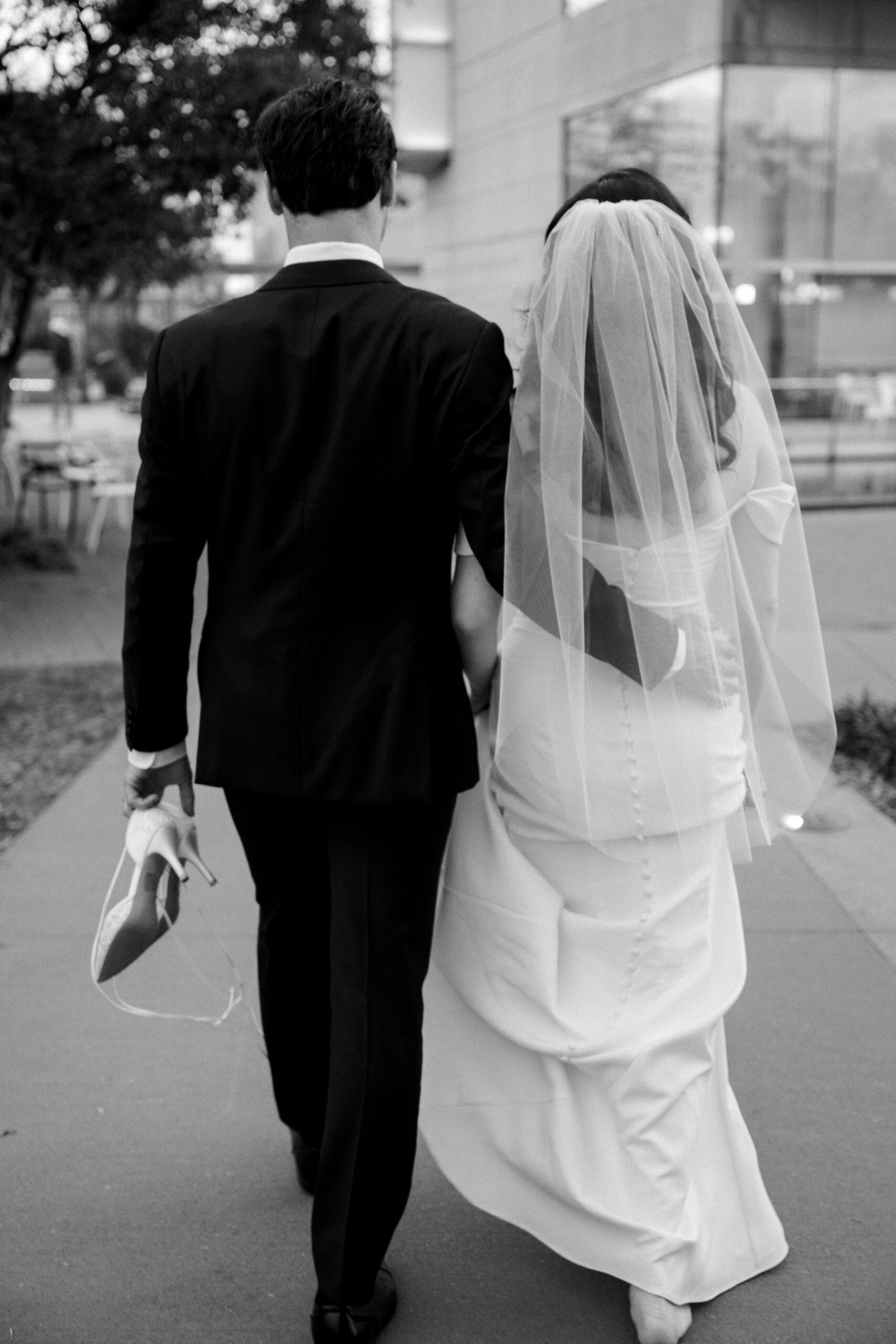 Black and white full body image of bride and groom walking away from camera, groom has one hand on bride's back and with his other hand is carrying her shoes