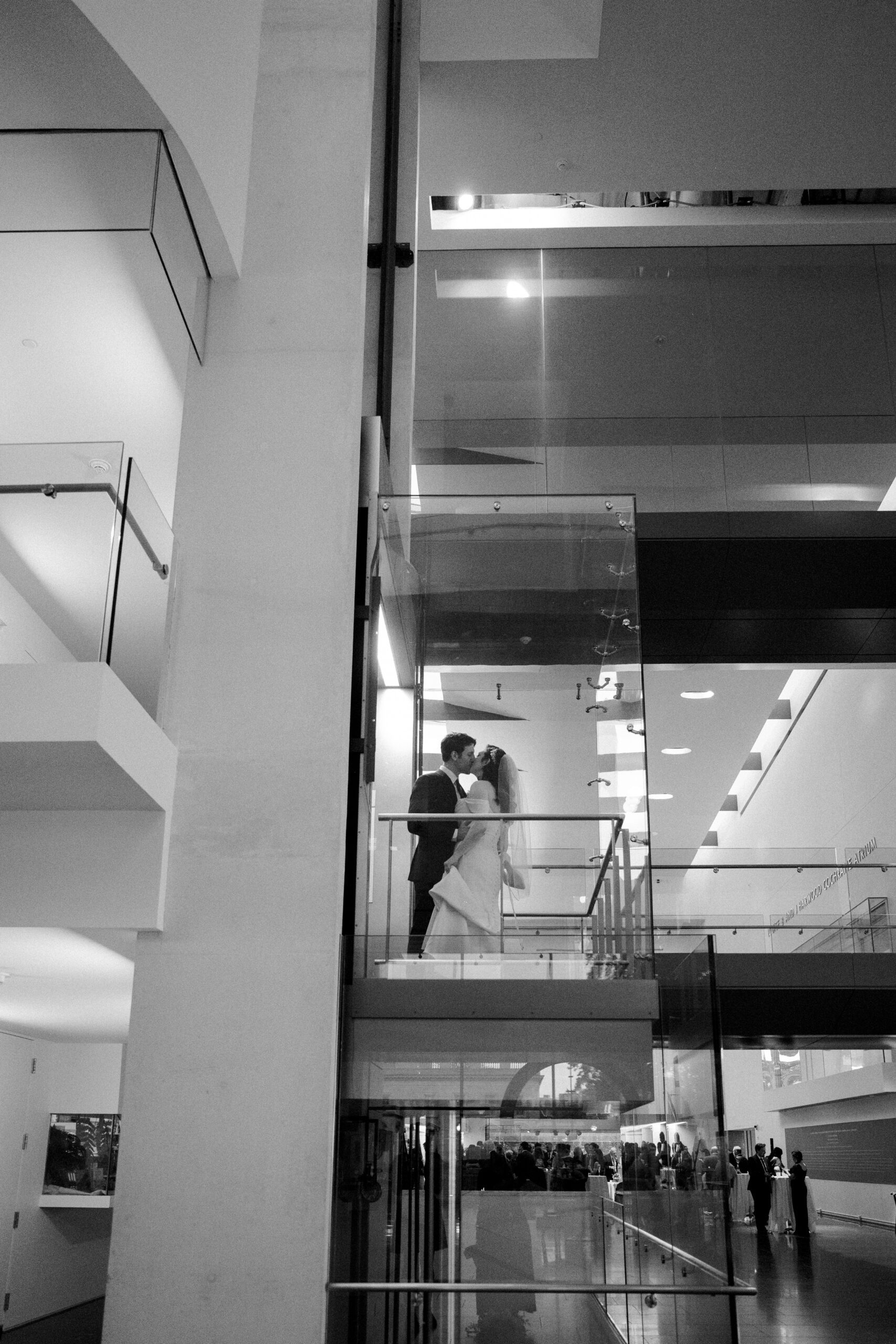 Distance photo of bride and groom sharing a kiss inside a clear elevator that is going up at vmfa