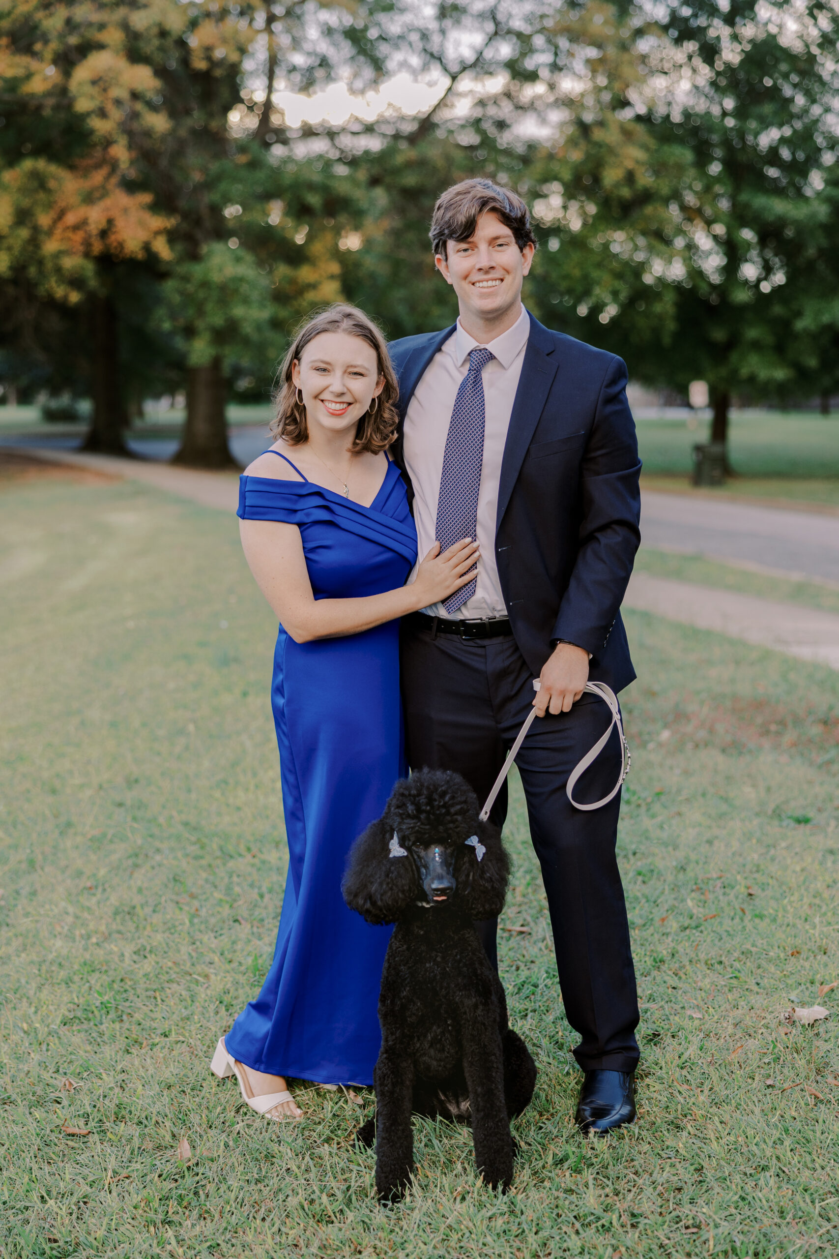 Woman in blue dress standing with her fiance in a suit, and their black poodle at byrd park