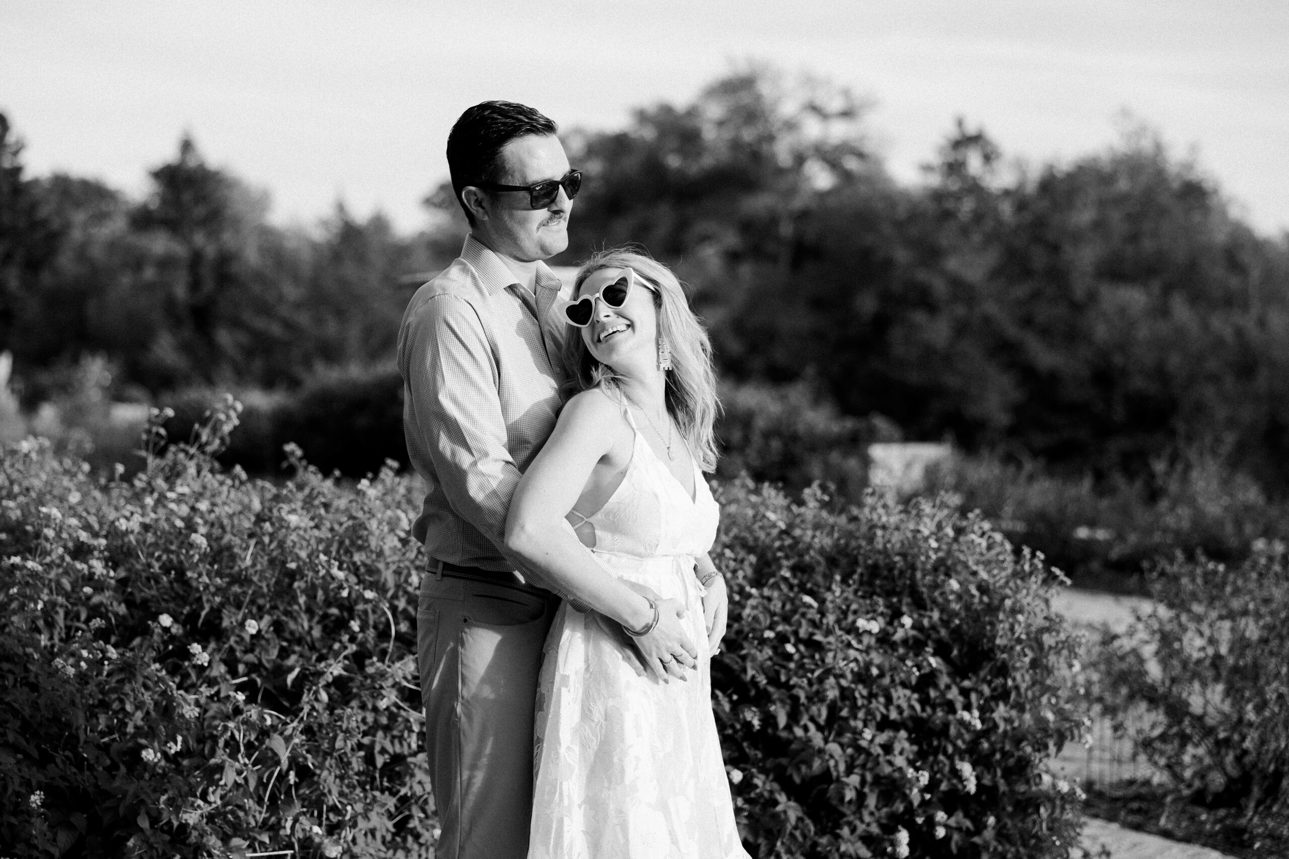 black and white photo of man wearing sunglasses hugging woman from behind, him facing the sun and her looking back at him. Woman is wearing heart shaped sunglasses