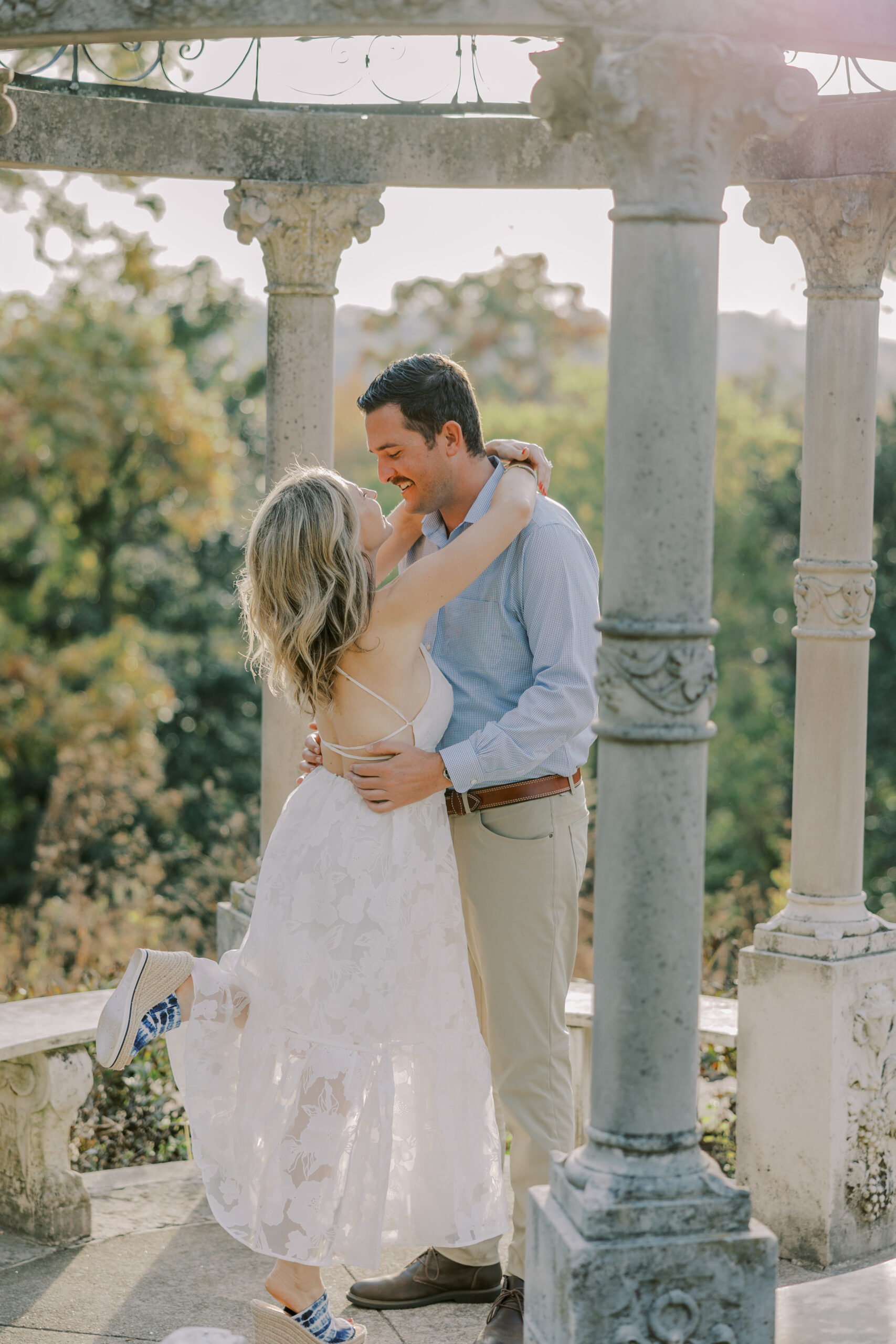 full length image of woman wearing white dress, arms wrapped around her fiance's neck as they look at each other in a stone gazebo