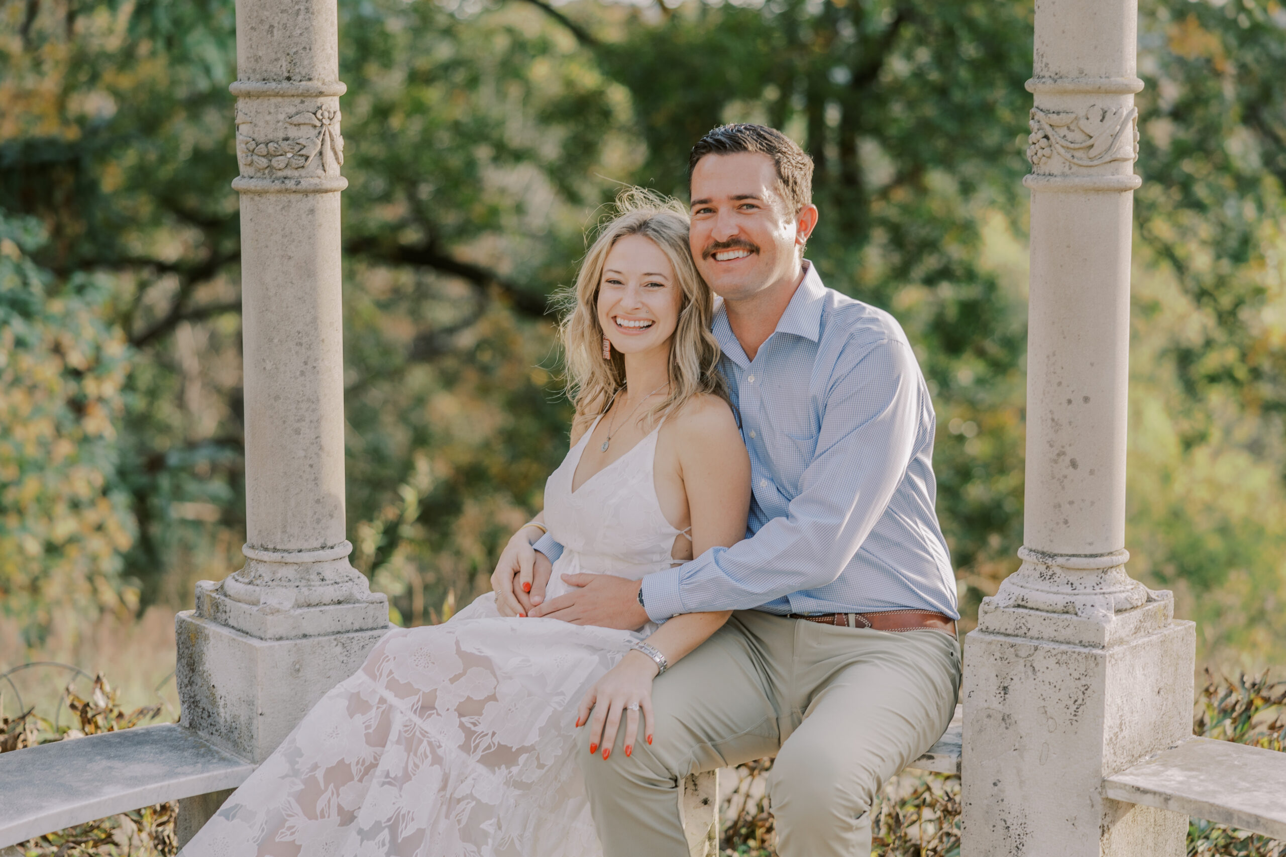 Woman in white dress and man in blue dress shirt and light brown dress pants sit on a stone bench at their maymont fall engagement session