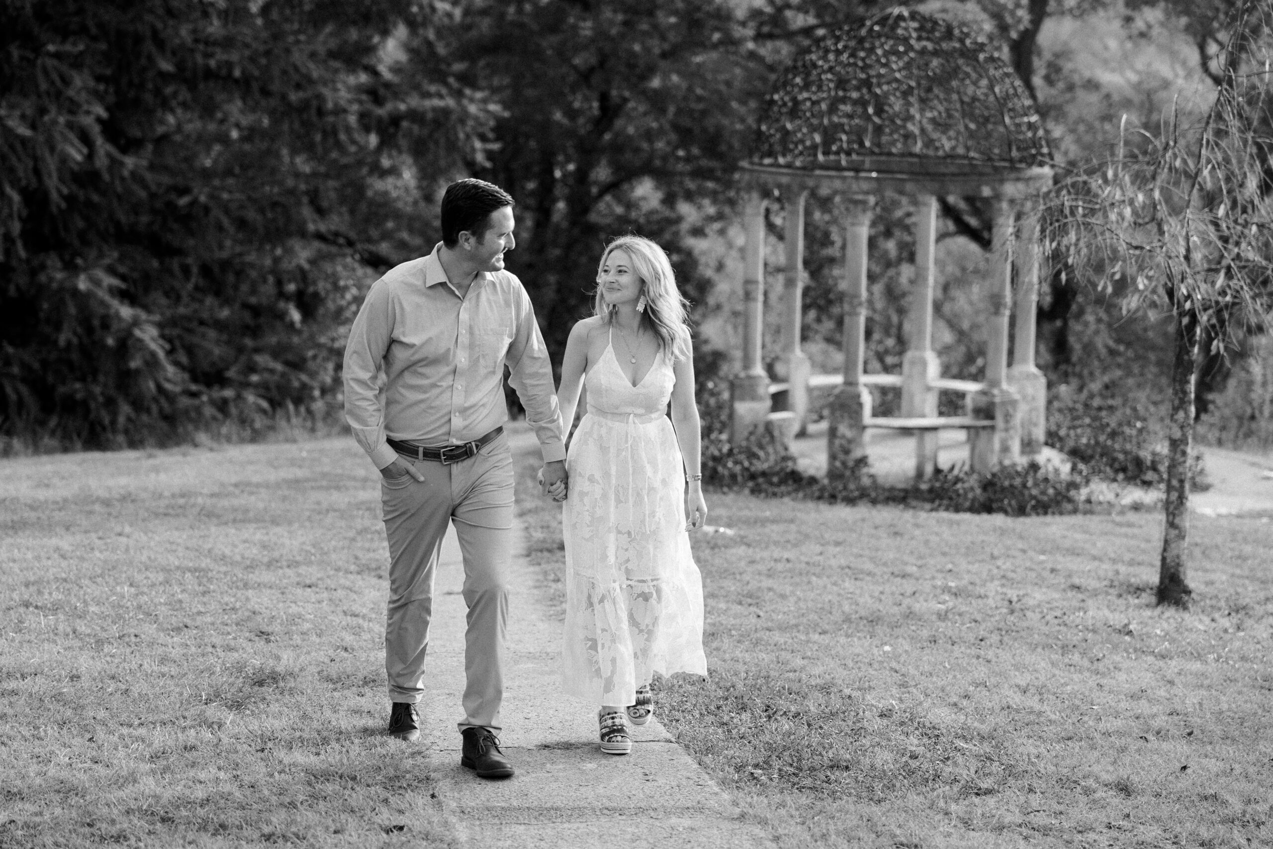 black and white image of couple walking on a small sidewalk with trees and a stone gazebo behind them at maymont