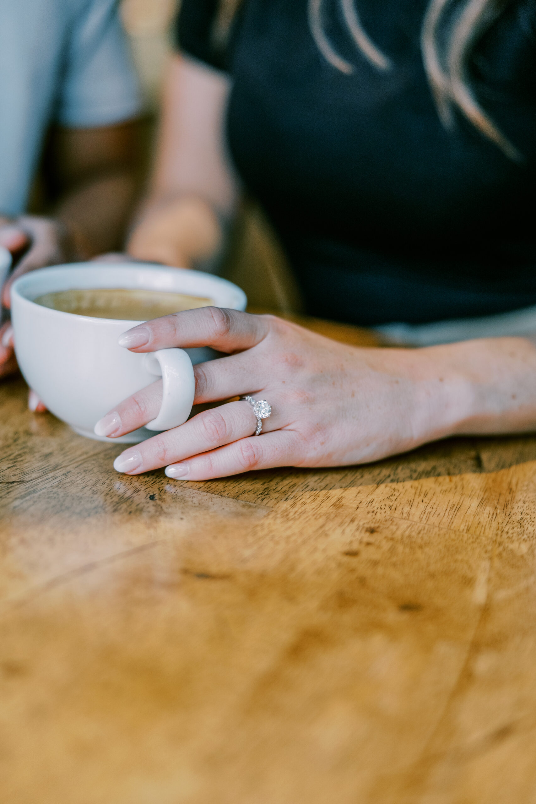 Detail photo of woman's hand on her coffee cup with her left hand, she has a diamond engagement ring on her finger