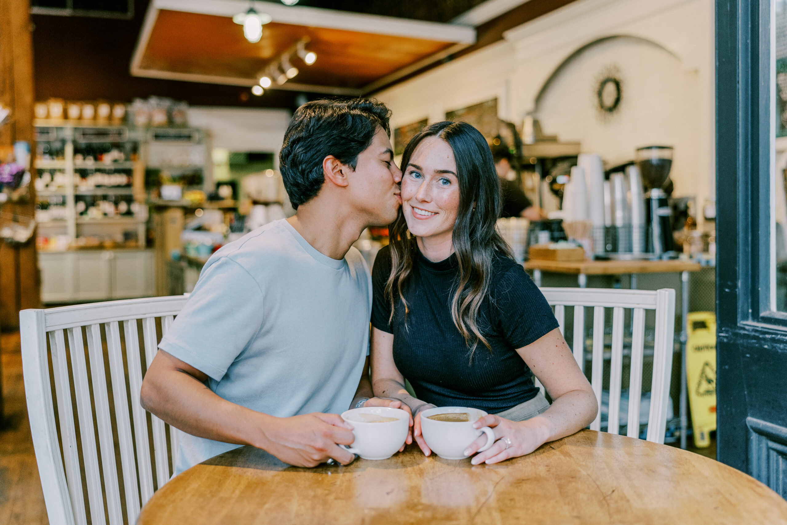 Man and woman sitting at table, man kisses woman on cheek as she looks at camera smiling