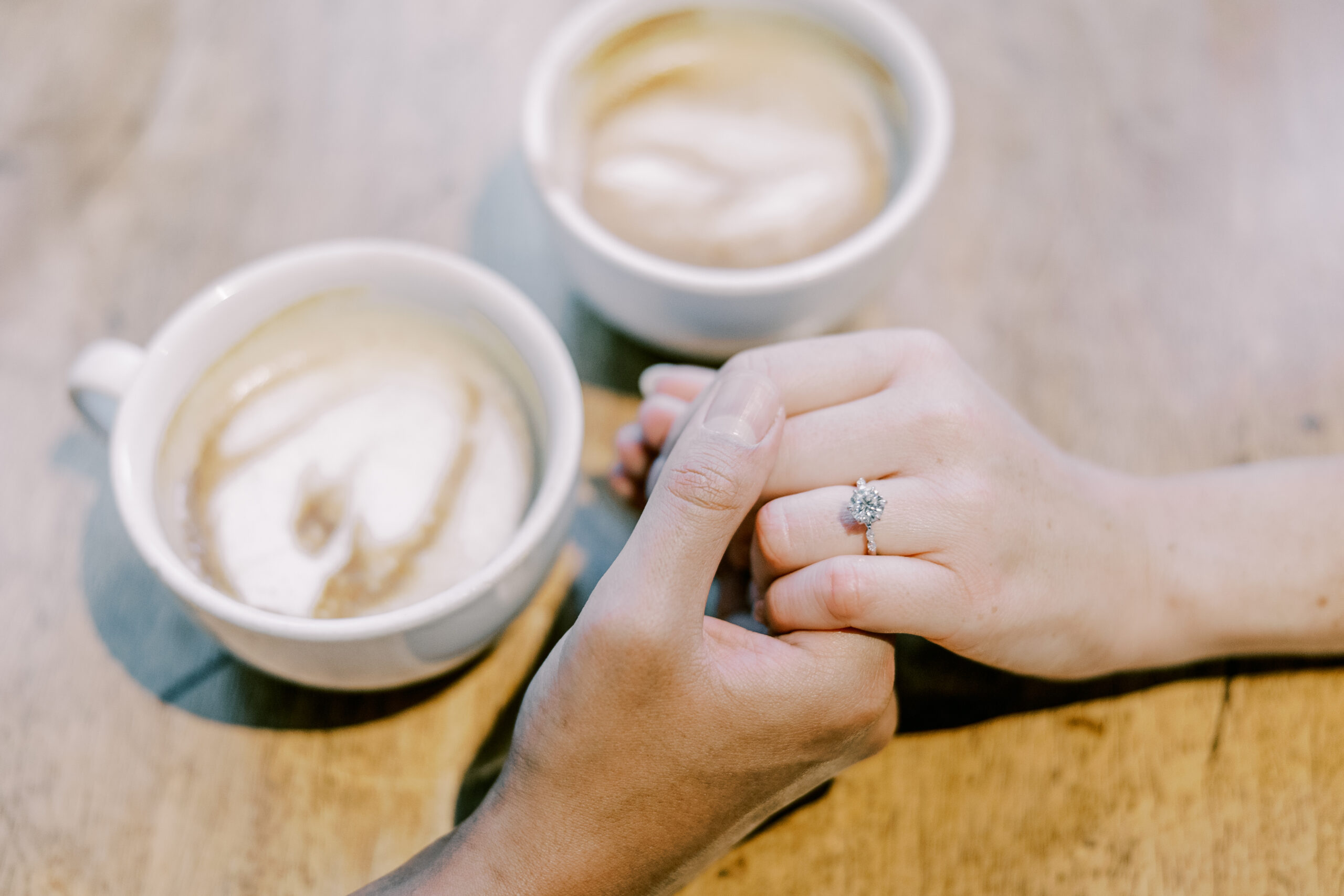 Close up image of man holding woman's hand showing off her engagement ring, two coffee mugs sit on the table beside them.