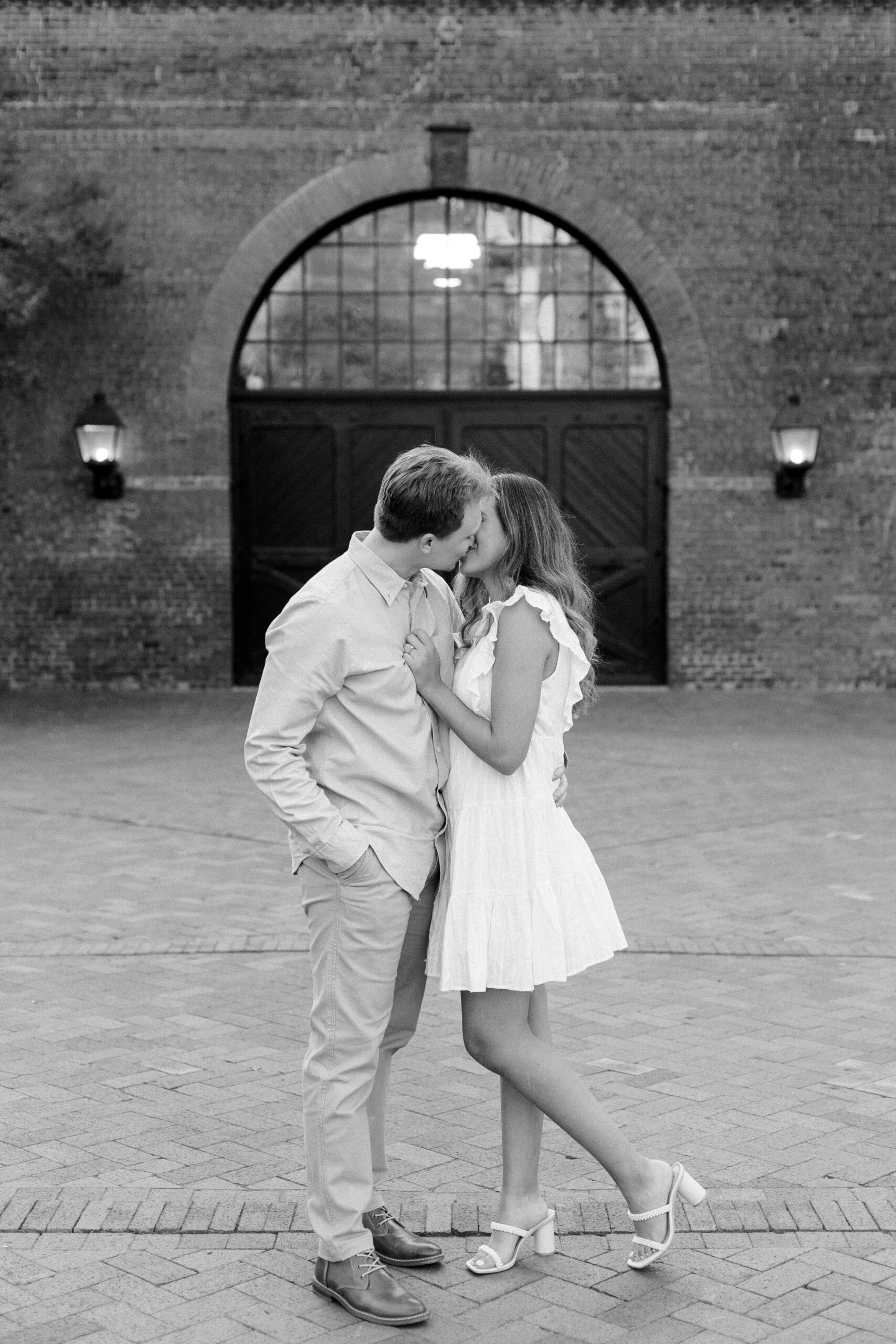 Black and white full length photo of couple kissing in front of brick wall with arched doors and windows behind them