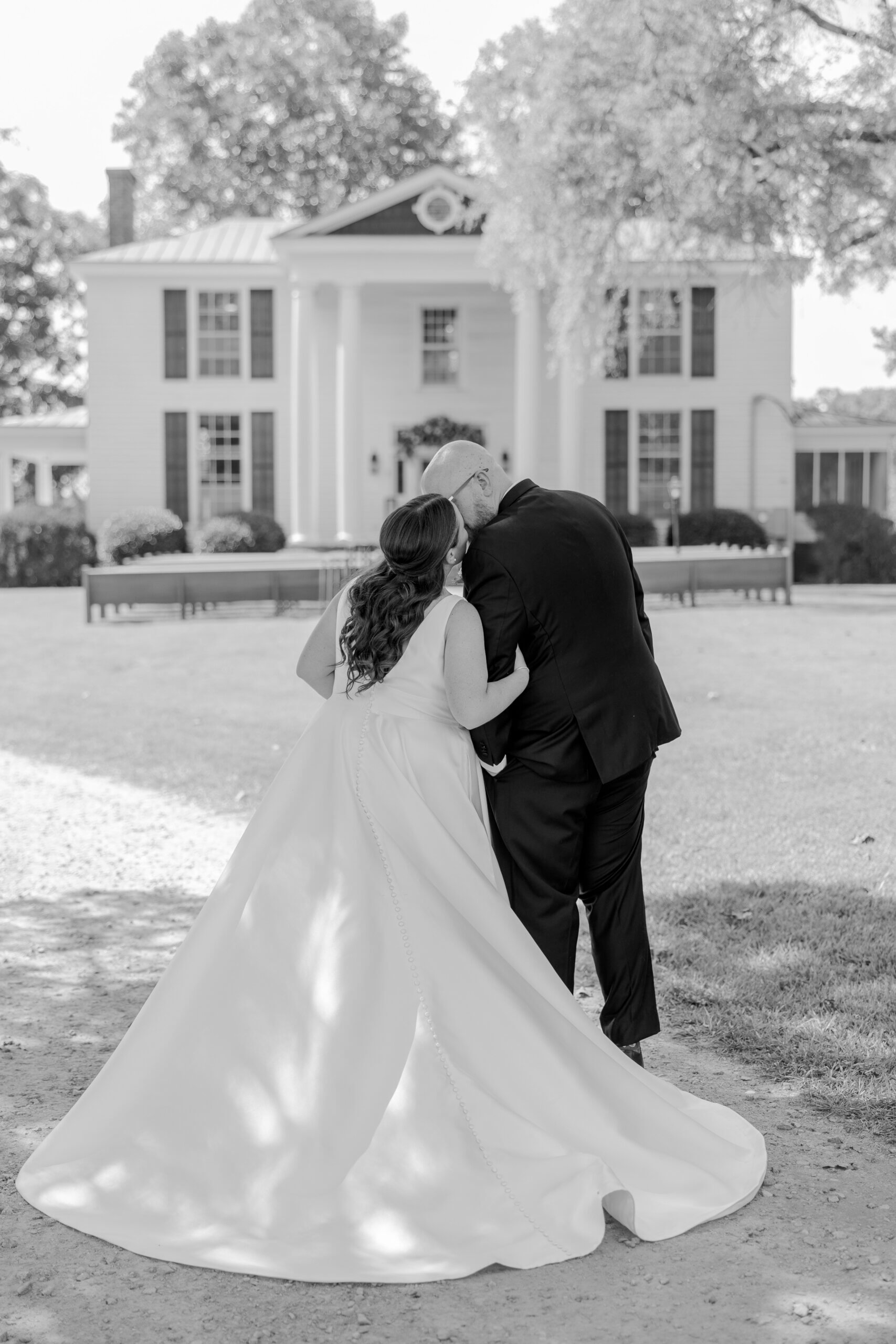 Black and white photo of bride and groom sharing a kiss, large white family famhouse in background