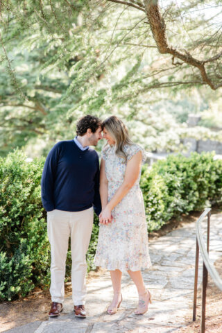 Woman in a white and colored floral dress holding hands with her fiance whos in light brown pants and a dark blue sweater touching noses smiling at one another