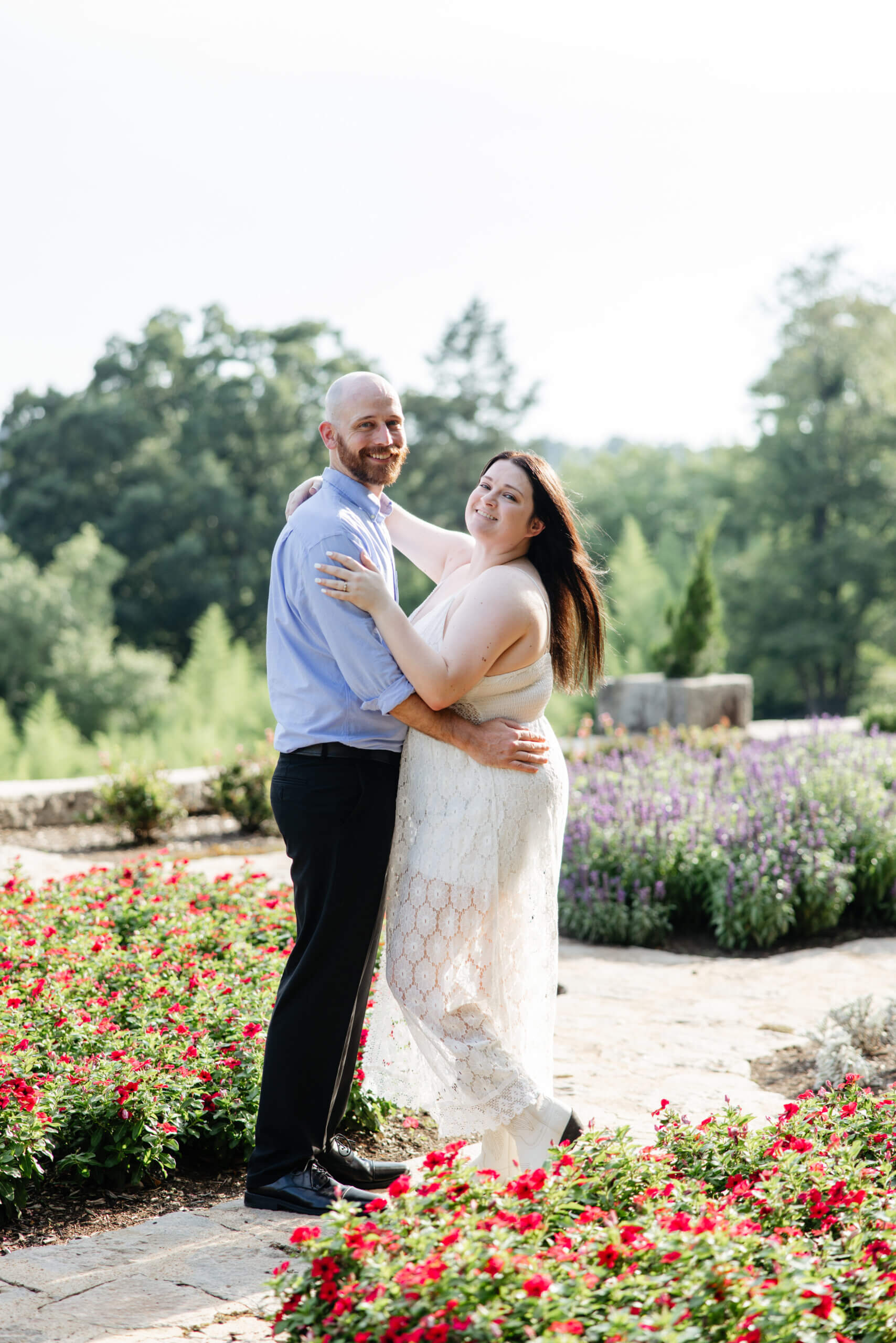 Man and woman; engaged couple, standing in the flower gardens at maymont park with their arms around one another smiling at camera