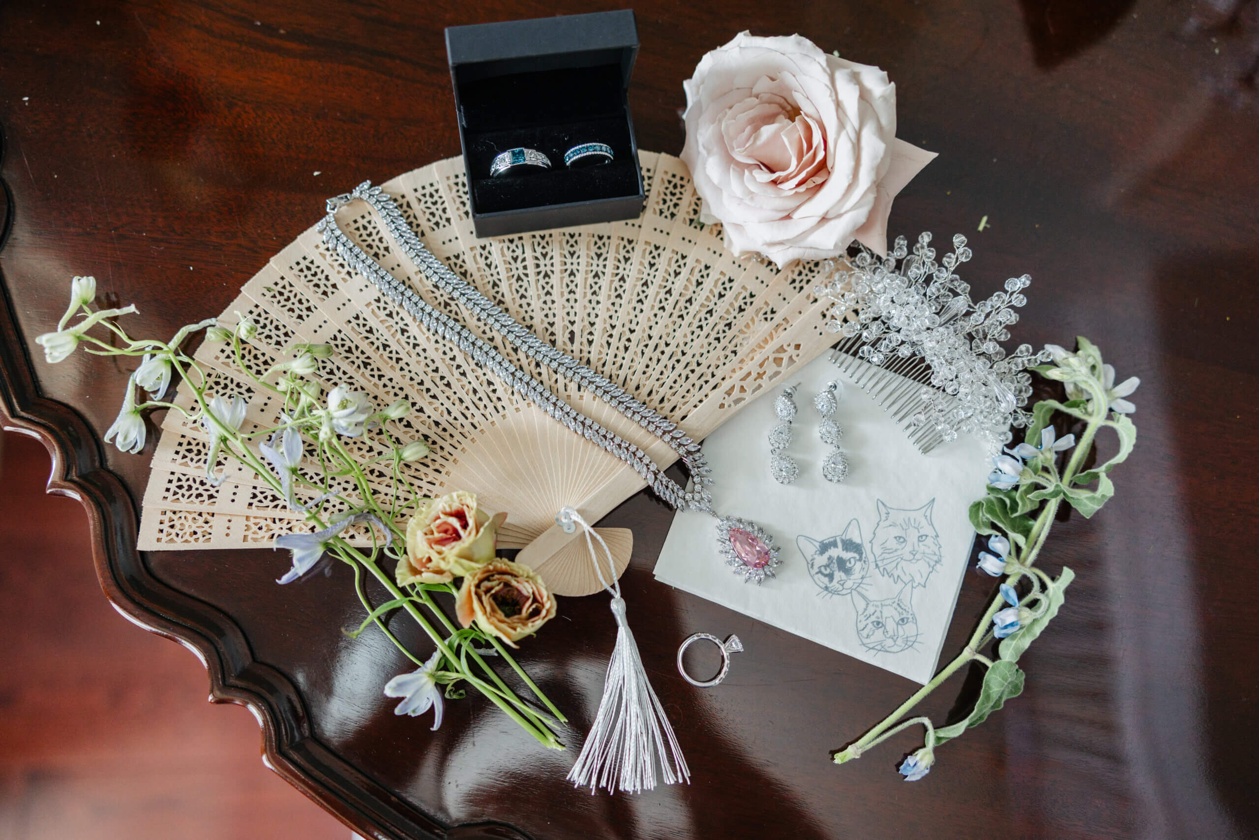 Private Residence Summer Wedding 1 Cream colored hand fan laying on table with bride and groom's rings, necklace, a napkin with their cats on it, and some florals