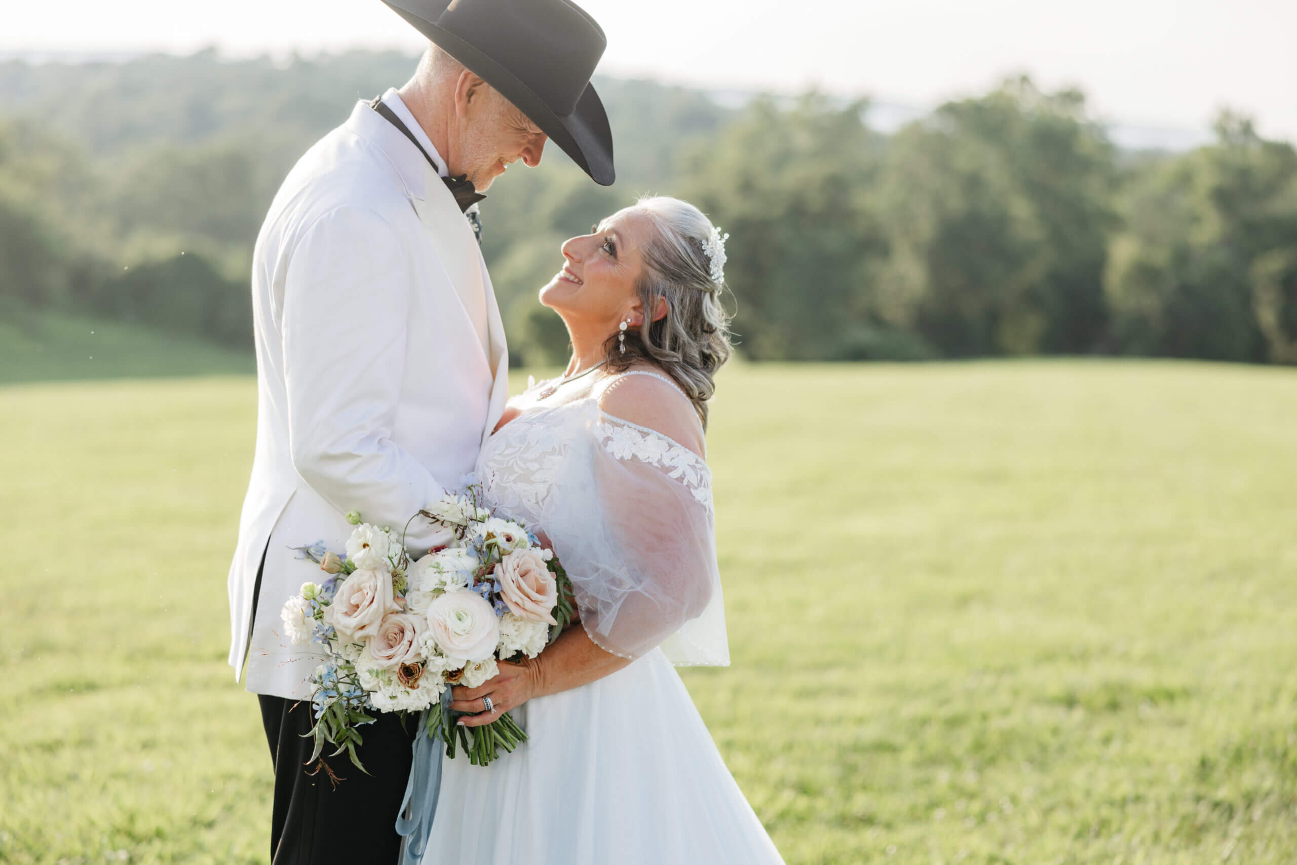 Private Residence Summer Wedding 24 Bride and groom smiling at one another as bride holds bouquet of off white, light pink/peach and light blue flowers, beautiful landscape in background