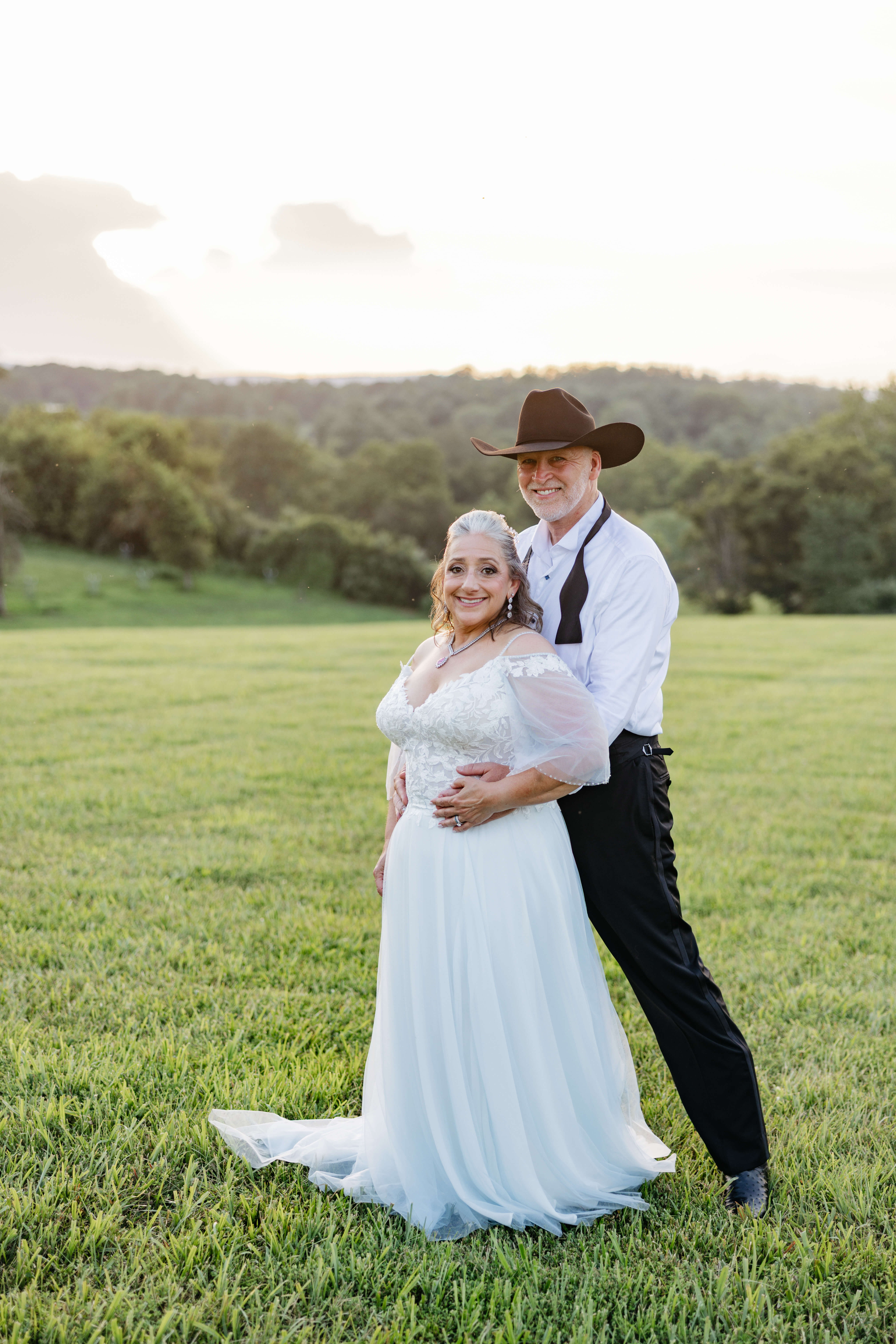 Private Residence Summer Wedding 34 Groom standing behind bride with his arms around her, both smiling at camera, sunset and green landscape in background
