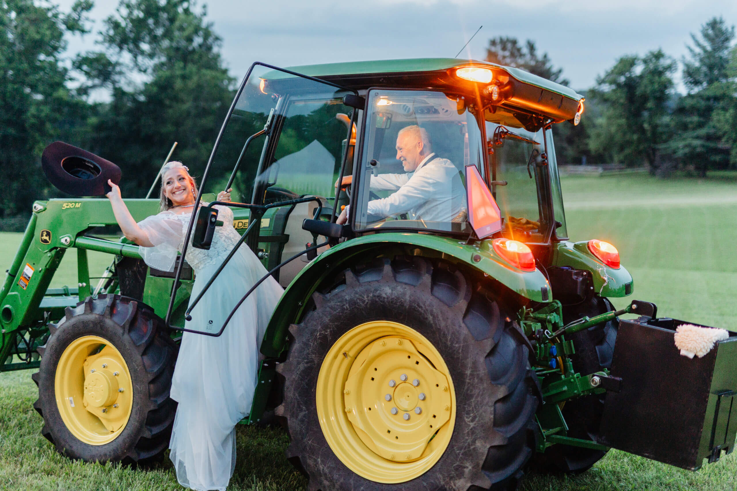 Private Residence Summer Wedding 40 Groom in tractor as bride climbs in, grabbing his hat, smiling back at guests