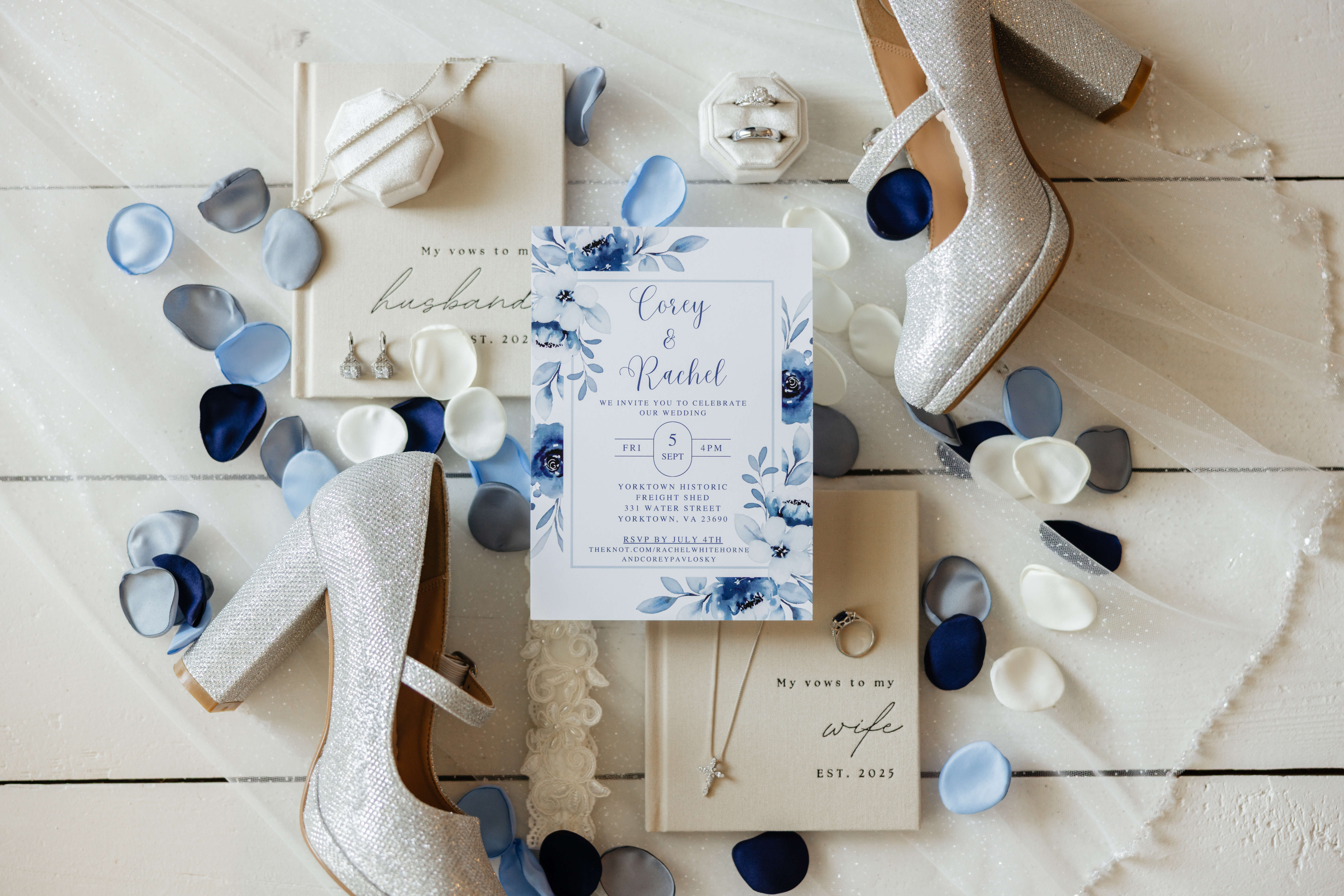 White and blue wedding invitation for corey and rachel sits with bride's sparkly silver shoes, off white vow books, bride's jewelry, and blue and white flower petals