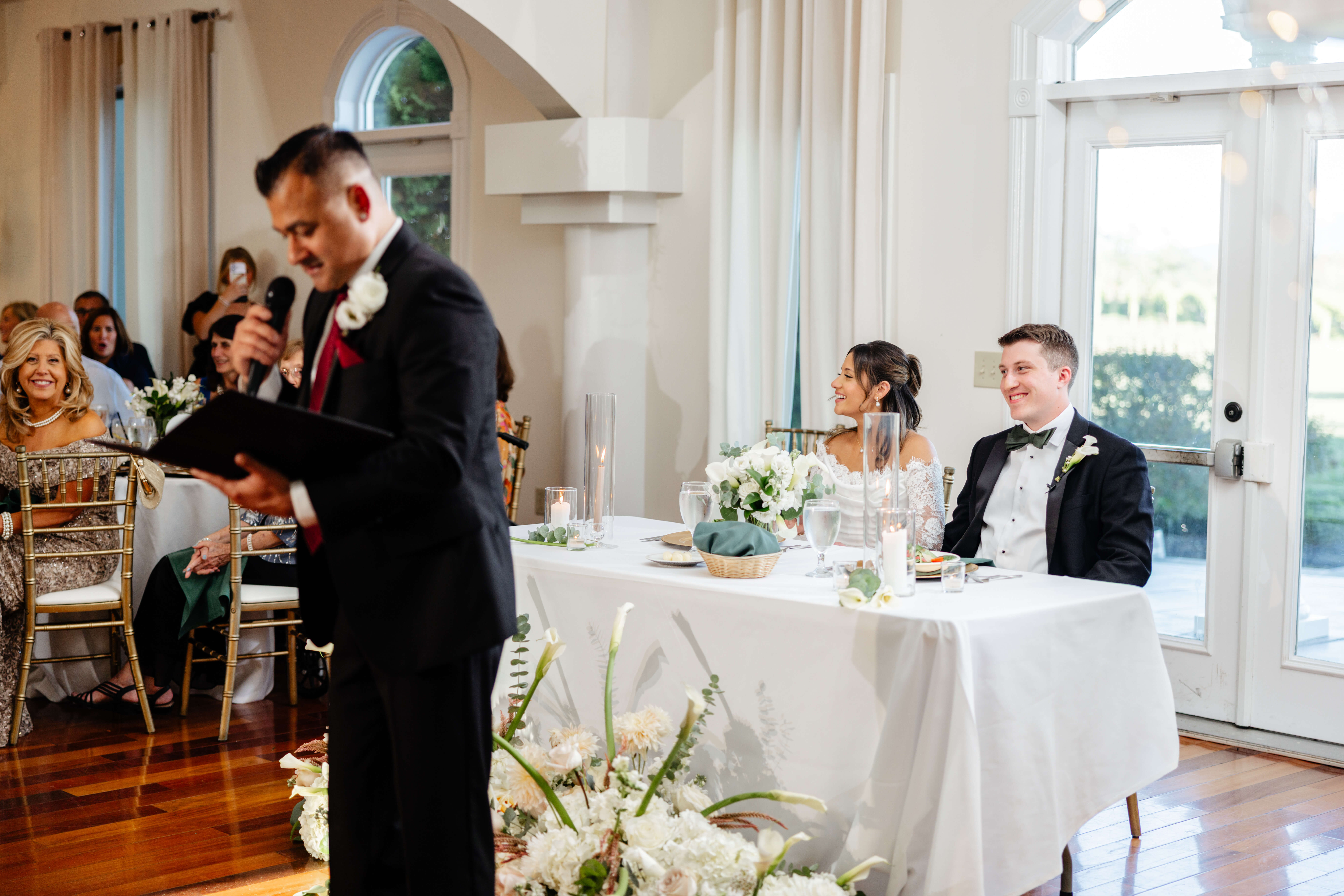 CrossKeys Vineyard Fall Wedding Photography 36 Bride and groom sit at sweetheart table smiling as bride's dad reads from folder into microphone