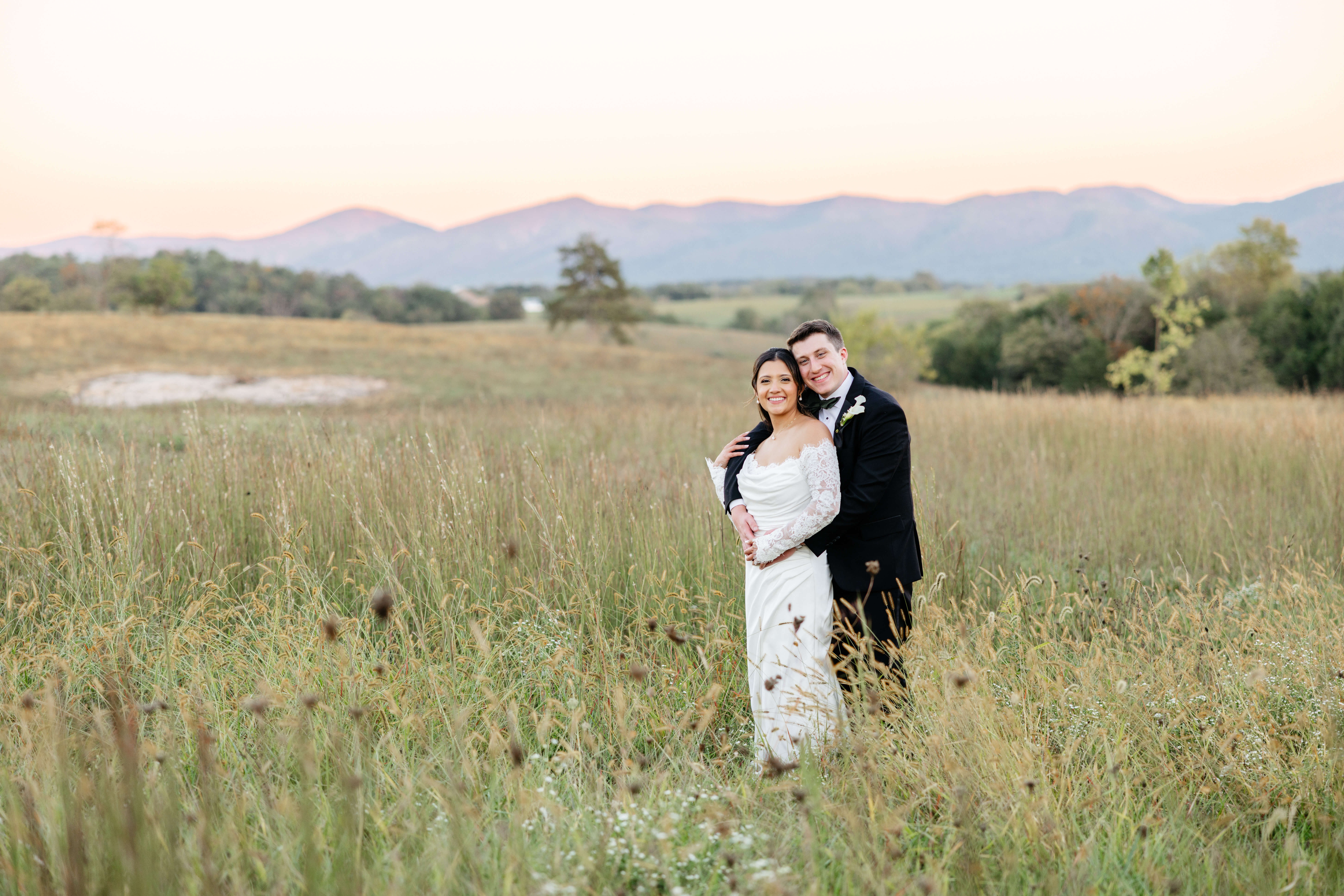 CrossKeys Vineyard Fall Wedding Photography 44 Bride and groom stand in tall grass at sunset with mountains in background both smiling at camera as groom hugs bride from behind