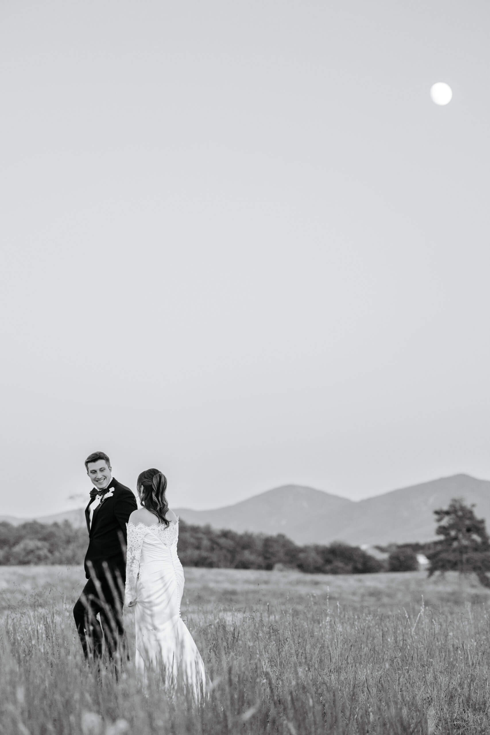 CrossKeys Vineyard Fall Wedding Photography 45 Black and white photo of bride and groom walking through long grassy area, mountains in background and the moon in the sky