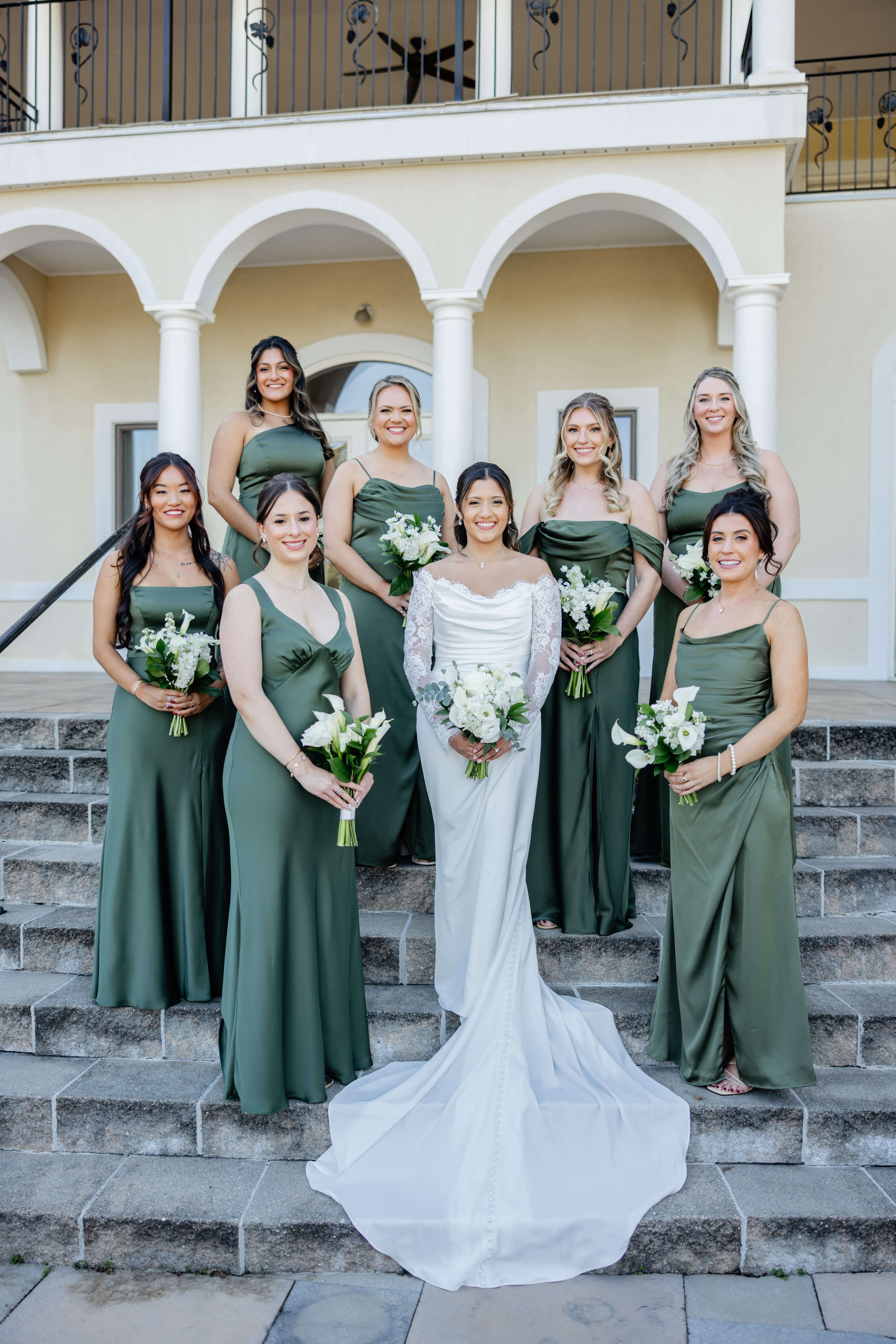 CrossKeys Vineyard Fall Wedding Photography 17 Bride in her wedding dress and her seven bridesmaids all in rich green colored bridesmaid dresses stand on grey stone outdoor stairs holding bouquets of white flowers