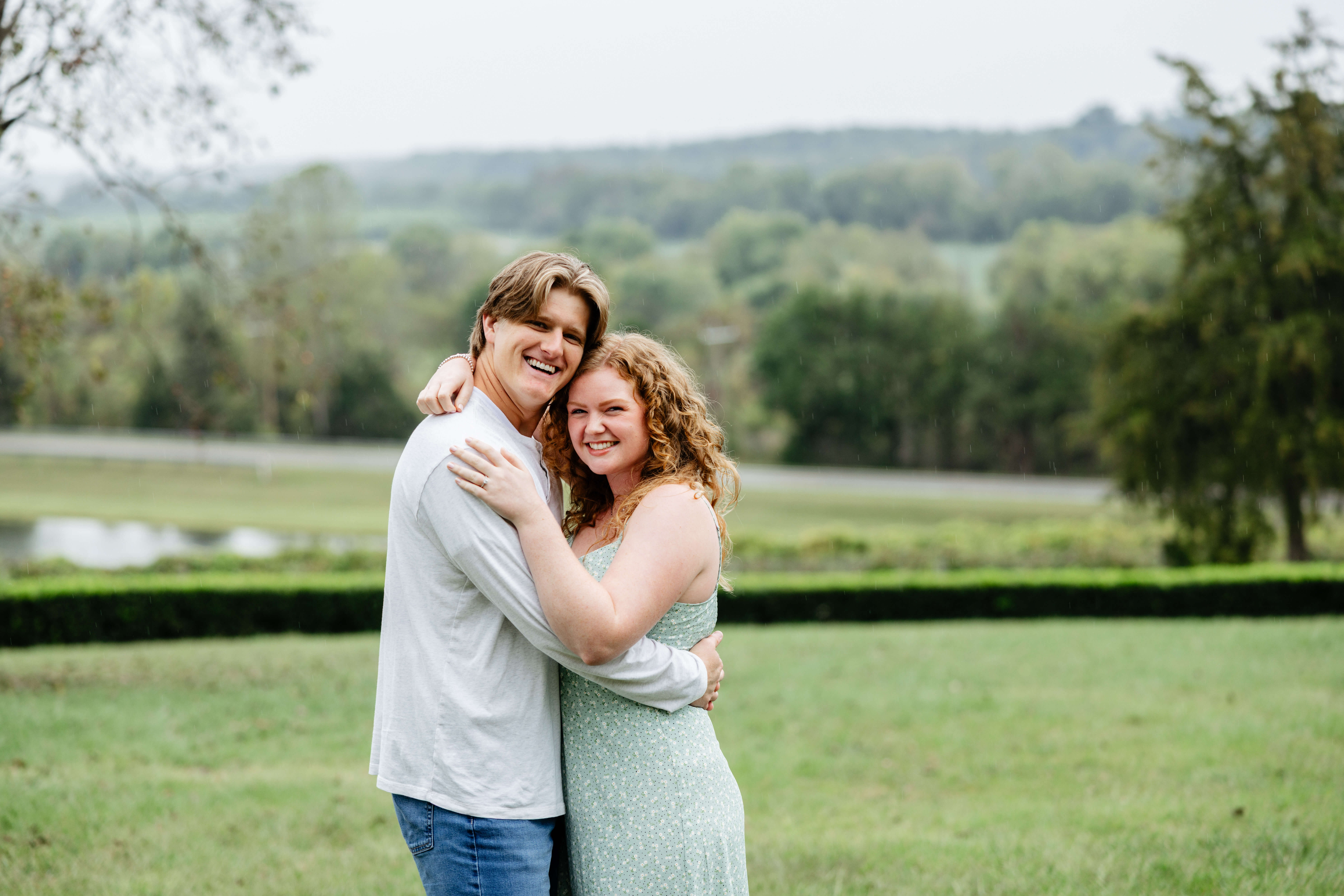 Rassawek Vineyards Fall Engagement Photography 1 Woman in light green dress hugging her fiance whos wearing a white long sleeve shirt and jeans, beautiful greenery landscape in background