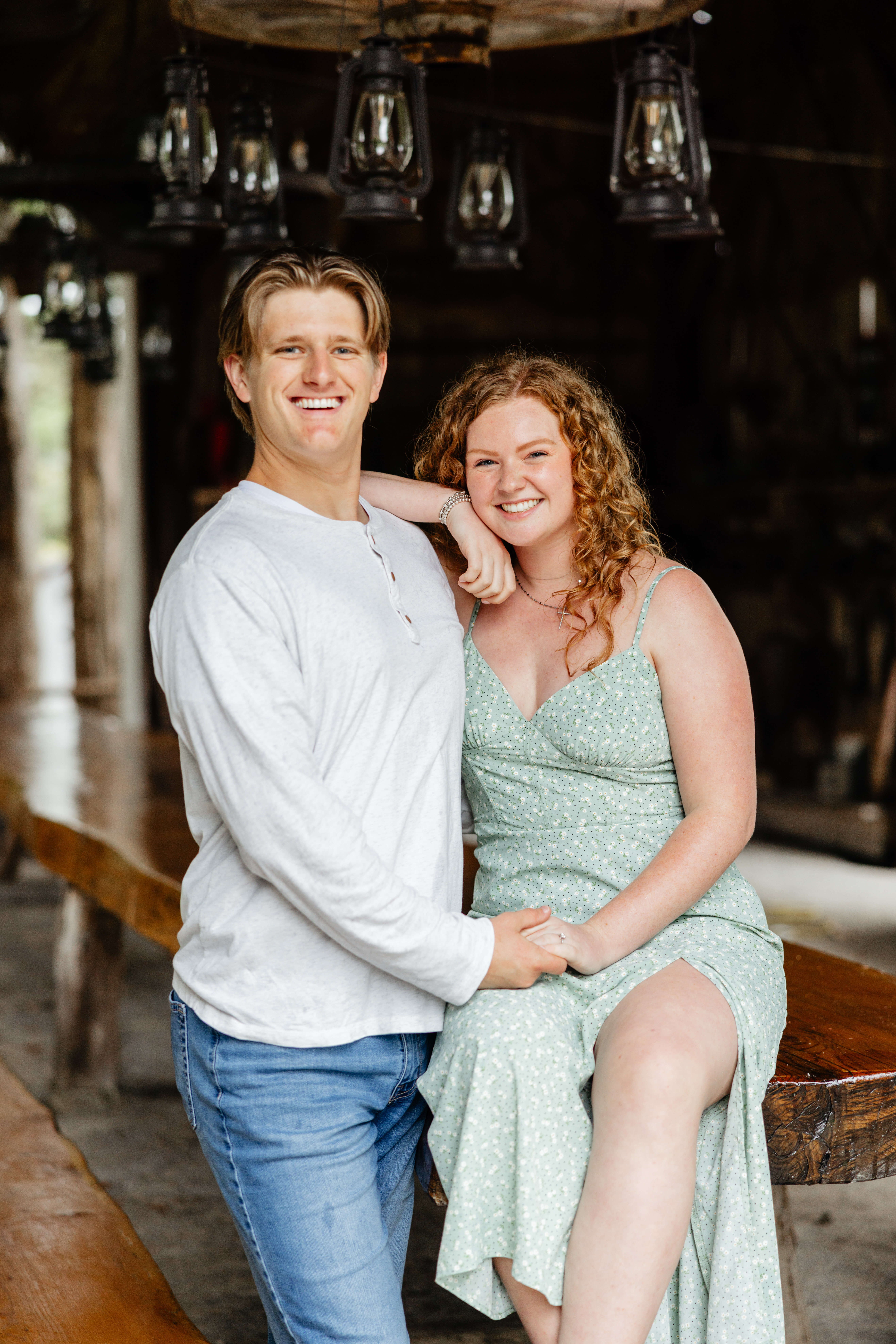 Rassawek Vineyards Fall Engagement Photography 6 Woman sitting on table in covered area at rassawek vineyards as her fiance stands beside her holding her hand smiling