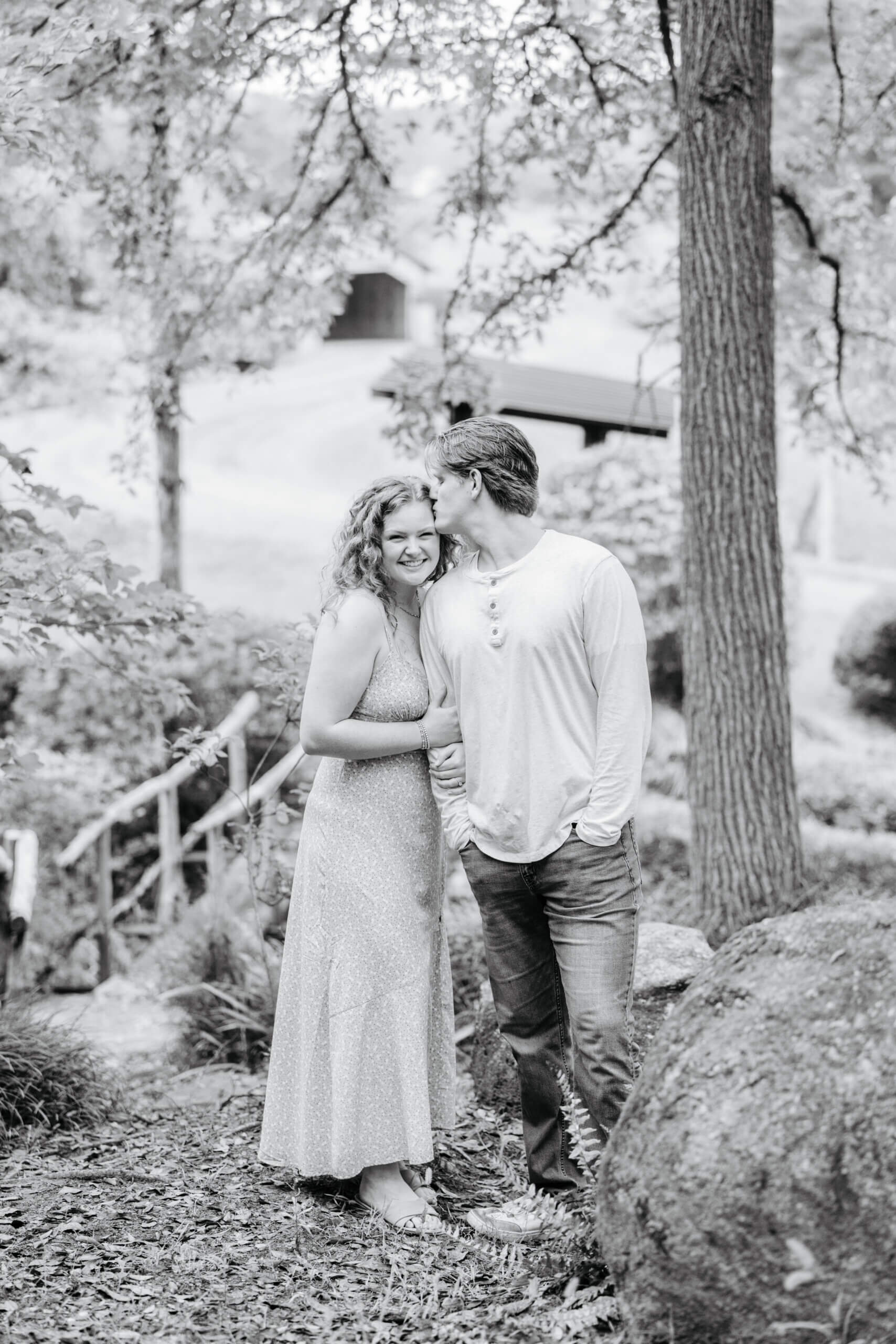 Rassawek Vineyards Fall Engagement Photography 8 Black and white photo of engaged couple standing in slight forest area as woman hugs man's arm and he kisses her head