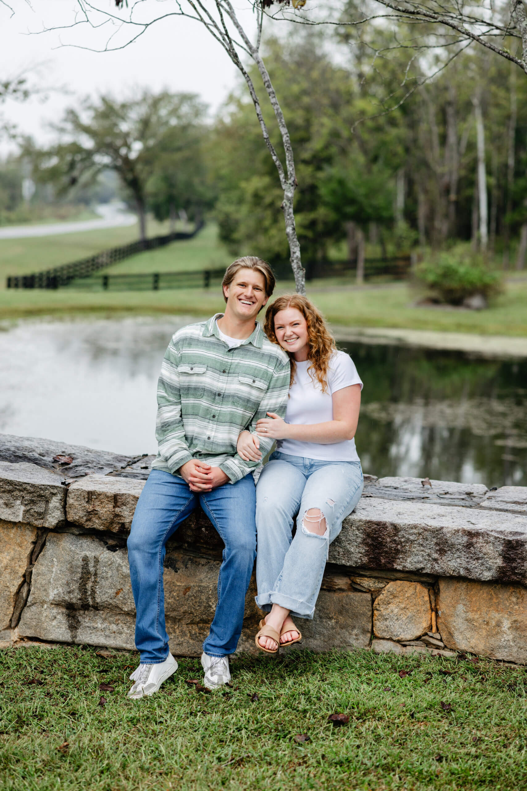 Rassawek Vineyards Fall Engagement Photography 15 Engaged couple sitting on stone short wall with pond behind them, woman hugging man's arm and both smile at camera