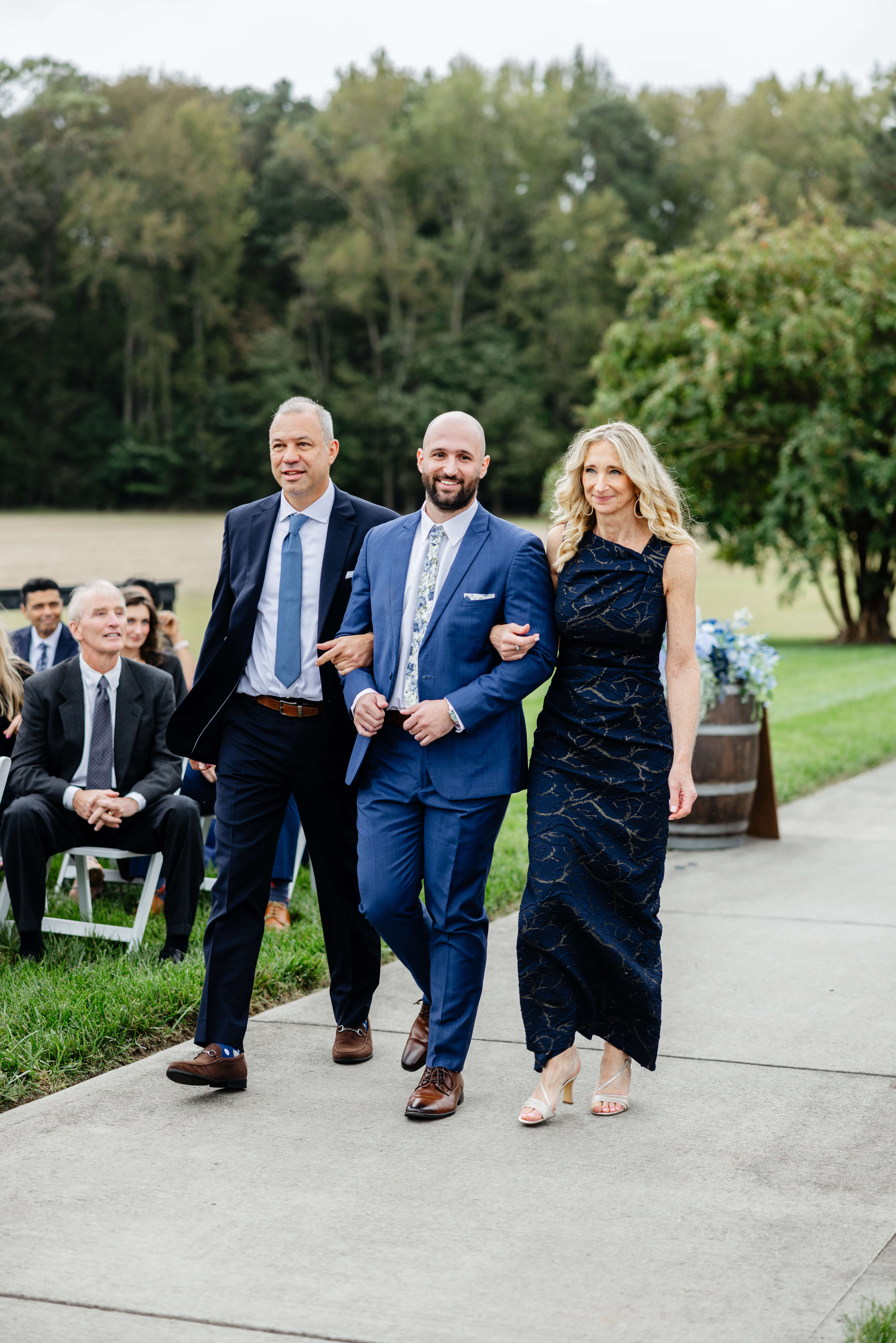 Barns of Kanak October Wedding 24 Groom walking down aisle arm in arm with both his mom and dad, mom is in a dark blue dress and dad is in a dark blue suit, groom in medium blue suit