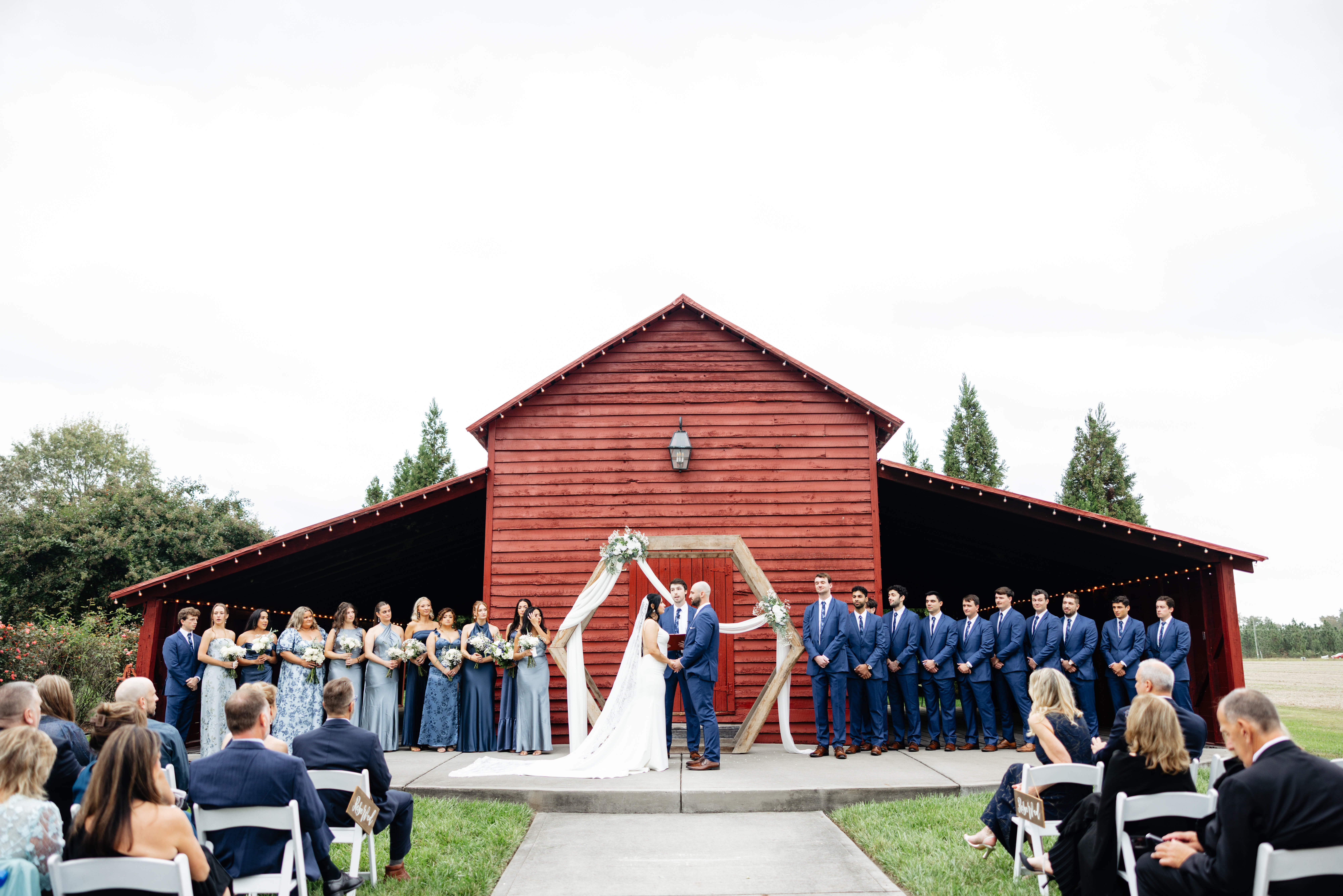 Barns of Kanak October Wedding 27 Red barn with bride, groom, officiant, and entire wedding party standing in front of it, bride and groom holding hands as officiant speaks